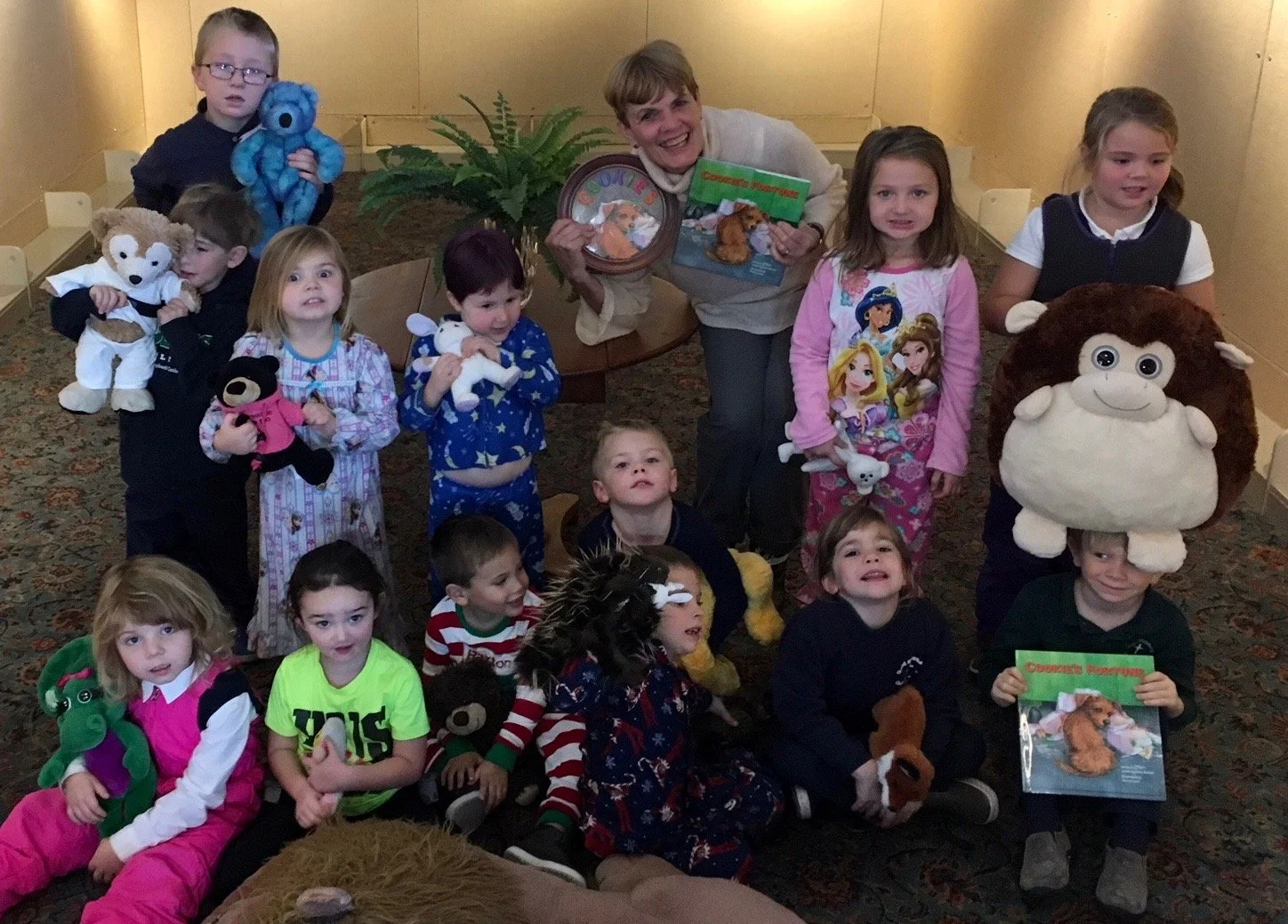 A group of children and an adult woman pose with stuffed animals, books, and plush toys in an indoor setting. Some children are sitting, others standing, and the woman stands in the center holding a book and a bowl. The children are dressed in pajama