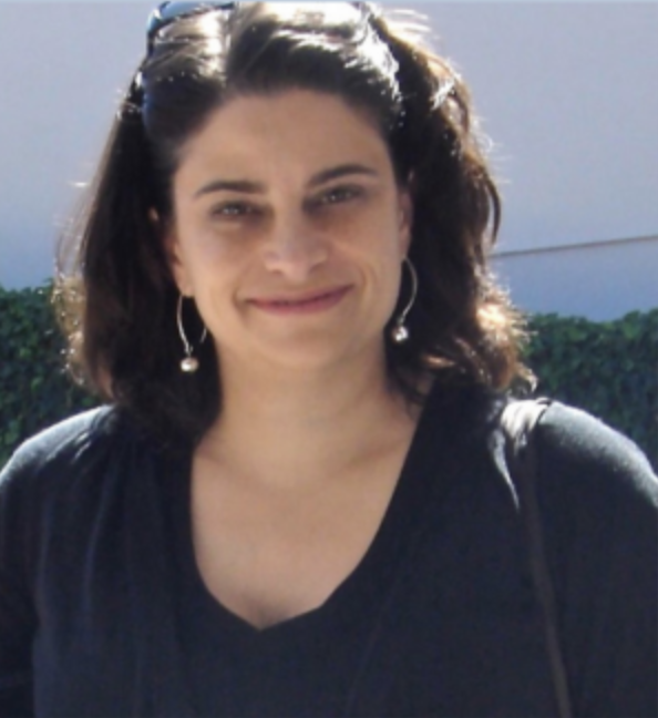 A woman with dark, curly hair and earrings, smiling, outdoors with a bright background.