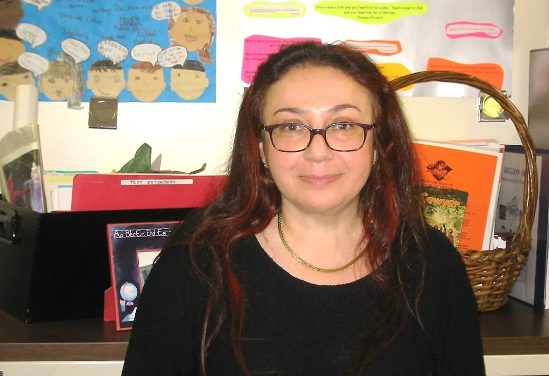 A woman with glasses and long red hair smiling while seated in front of a school or office desk with colorful posters and books behind her.