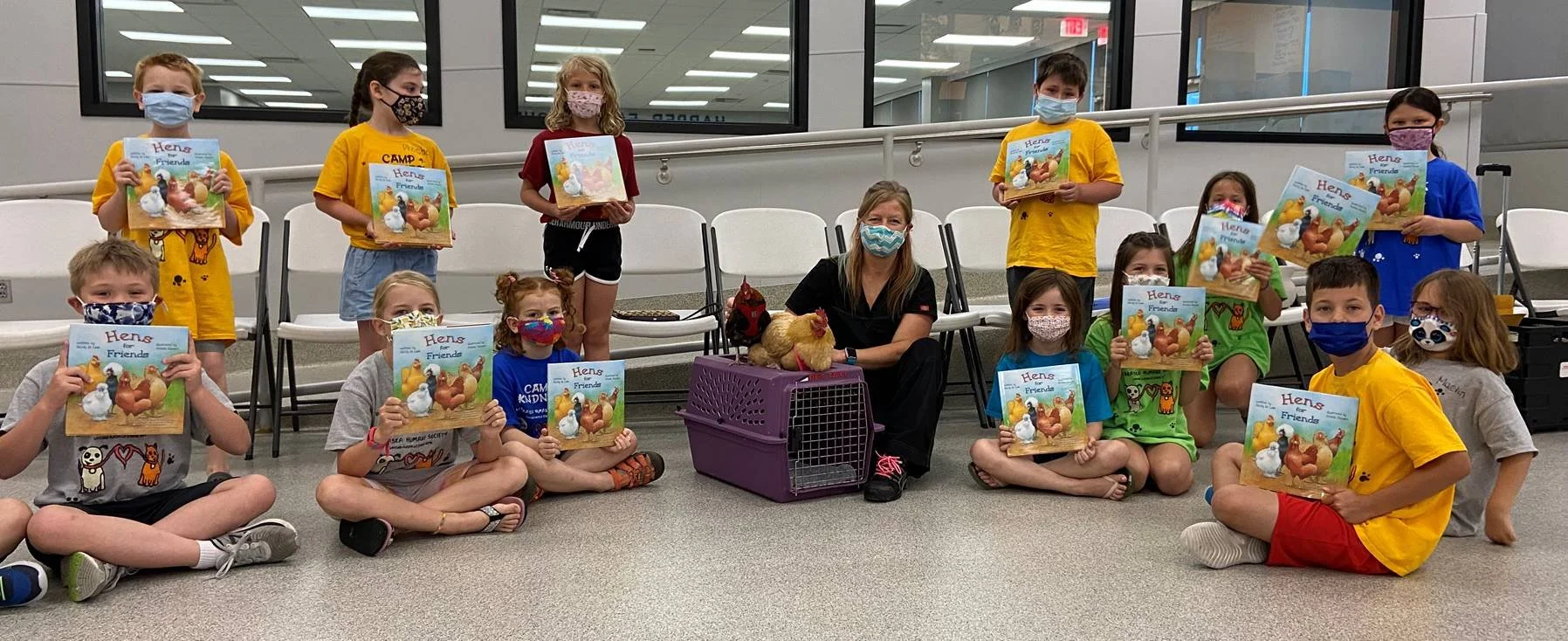Group of children sitting and standing with a woman, holding books titled 'Hens and Friends,' in an indoor setting with chairs, windows, and a purple pet carrier with a chicken inside.