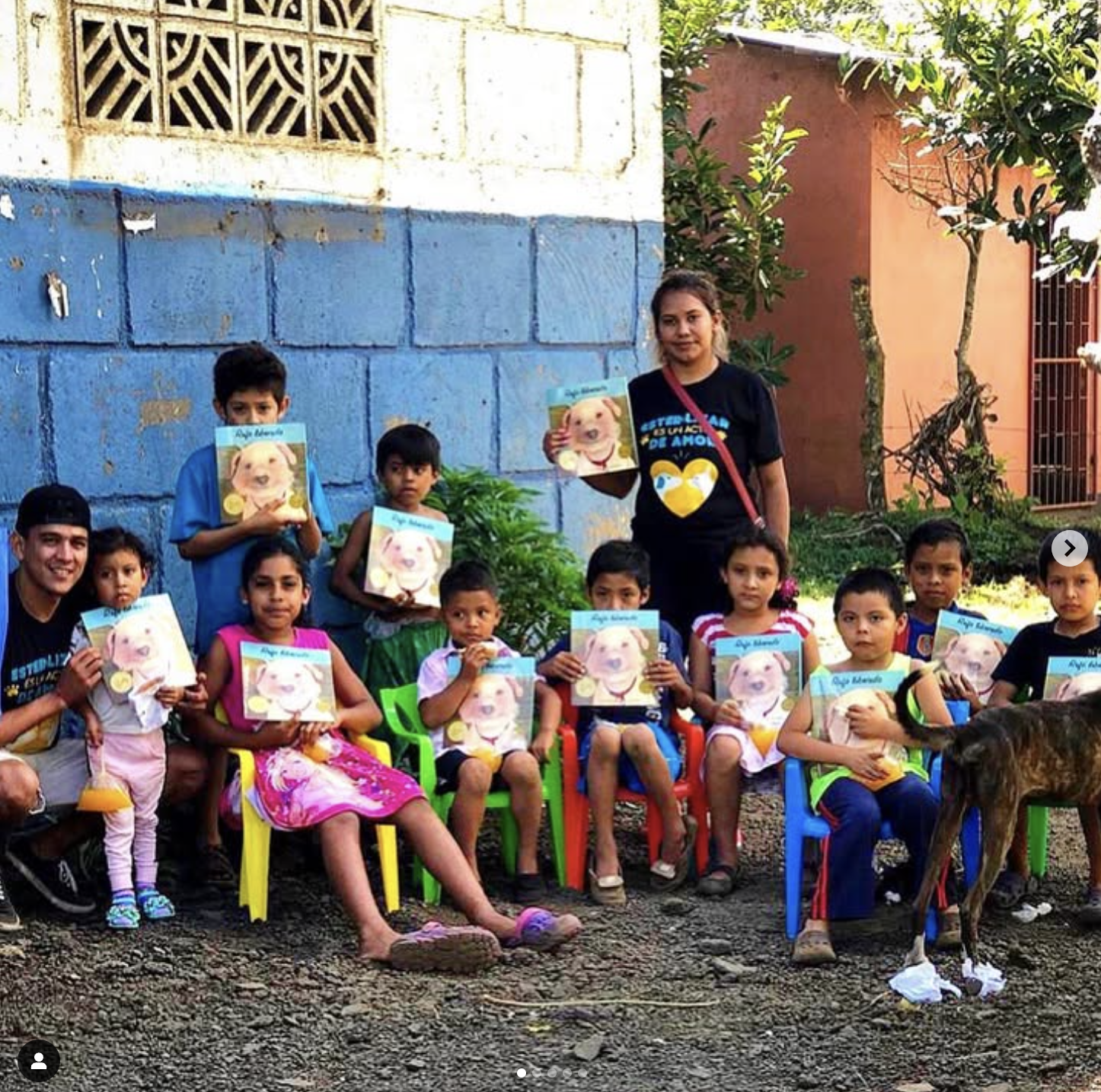 Group of children and two adults sitting and standing outdoors, holding books with a puppy on the cover, with a colorful background and a dog in front.