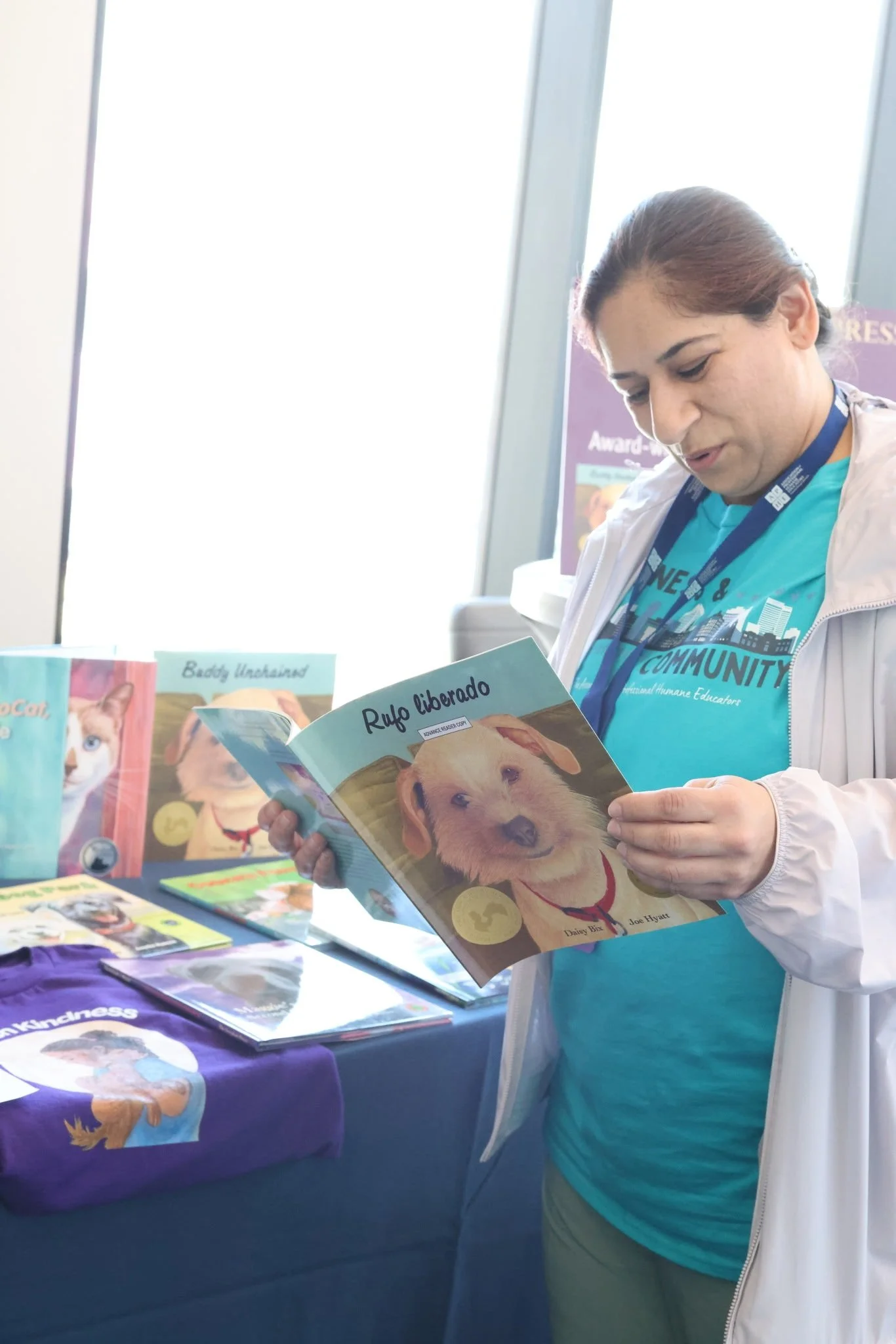 A woman wearing a blue shirt and white jacket is reading a booklet titled "Rufo liberado" featuring a golden retriever puppy on the cover. Behind her, a table displays pet-themed books and a purple shirt with a dog illustration.