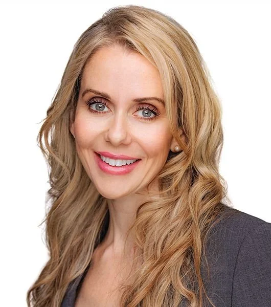 A woman with long blonde wavy hair smiling, wearing a dark blazer, against a white background.