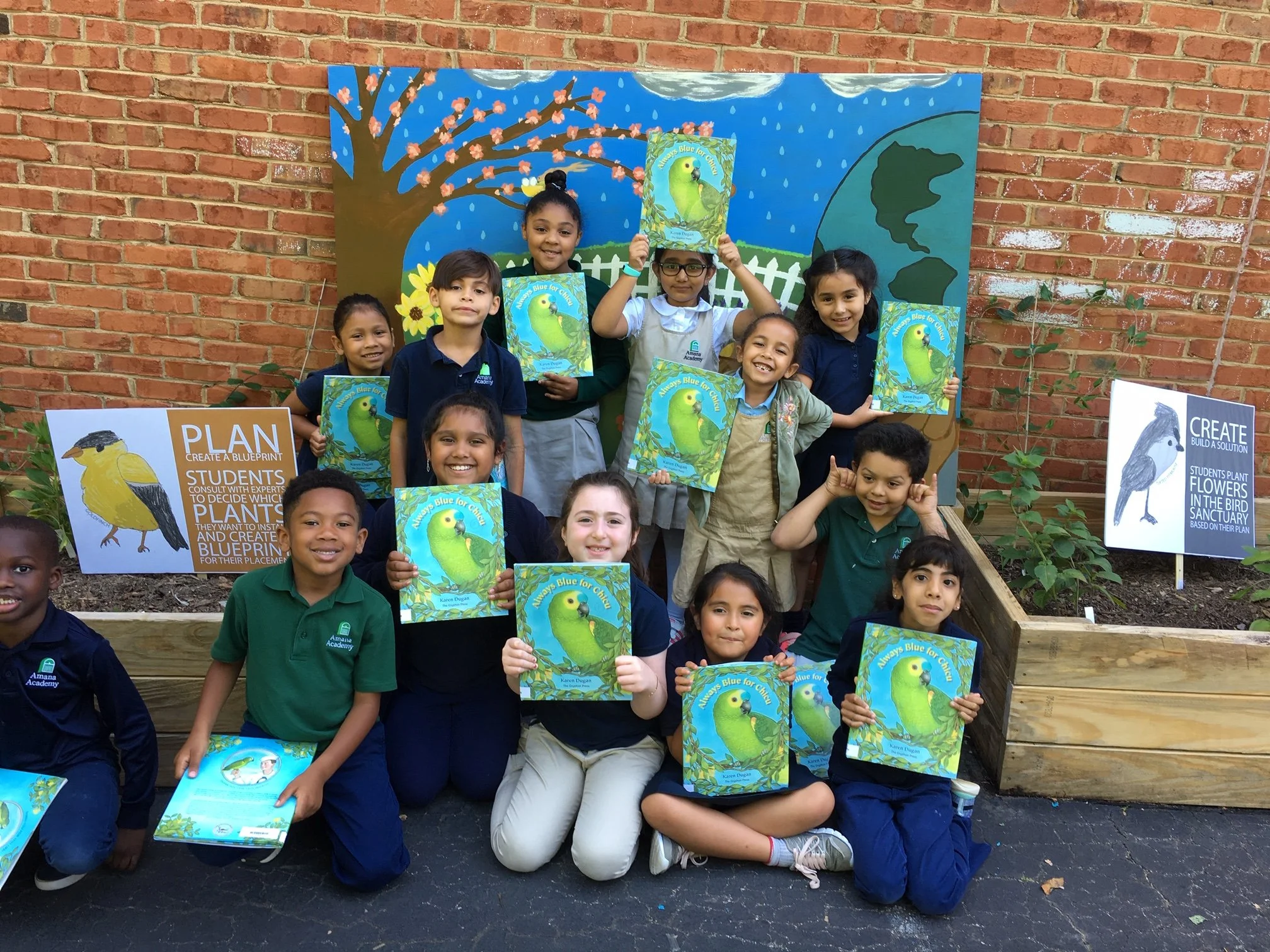 Group of children holding books about blue and green parrots, standing outdoors in front of a colorful mural featuring a tree with pink blossoms, a white picket fence, and a large green island. Signage about planting flowers and creating a blueprint 