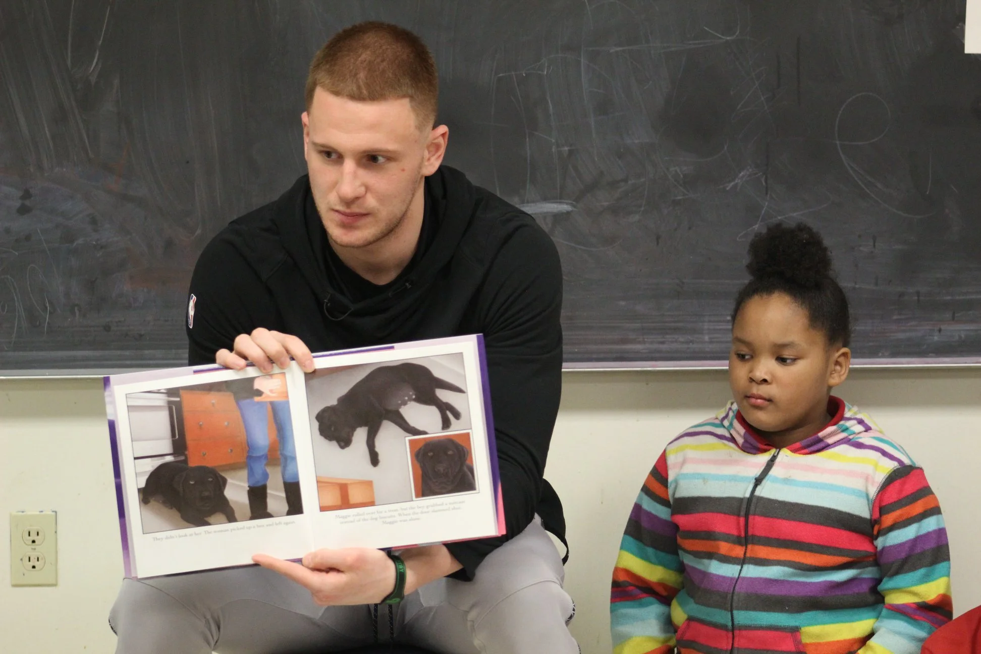 A man and a girl sitting in classroom, the man is holding a picture book with images of a dog, black Labrador puppy lying on the floor, standing, and a close-up of its face.