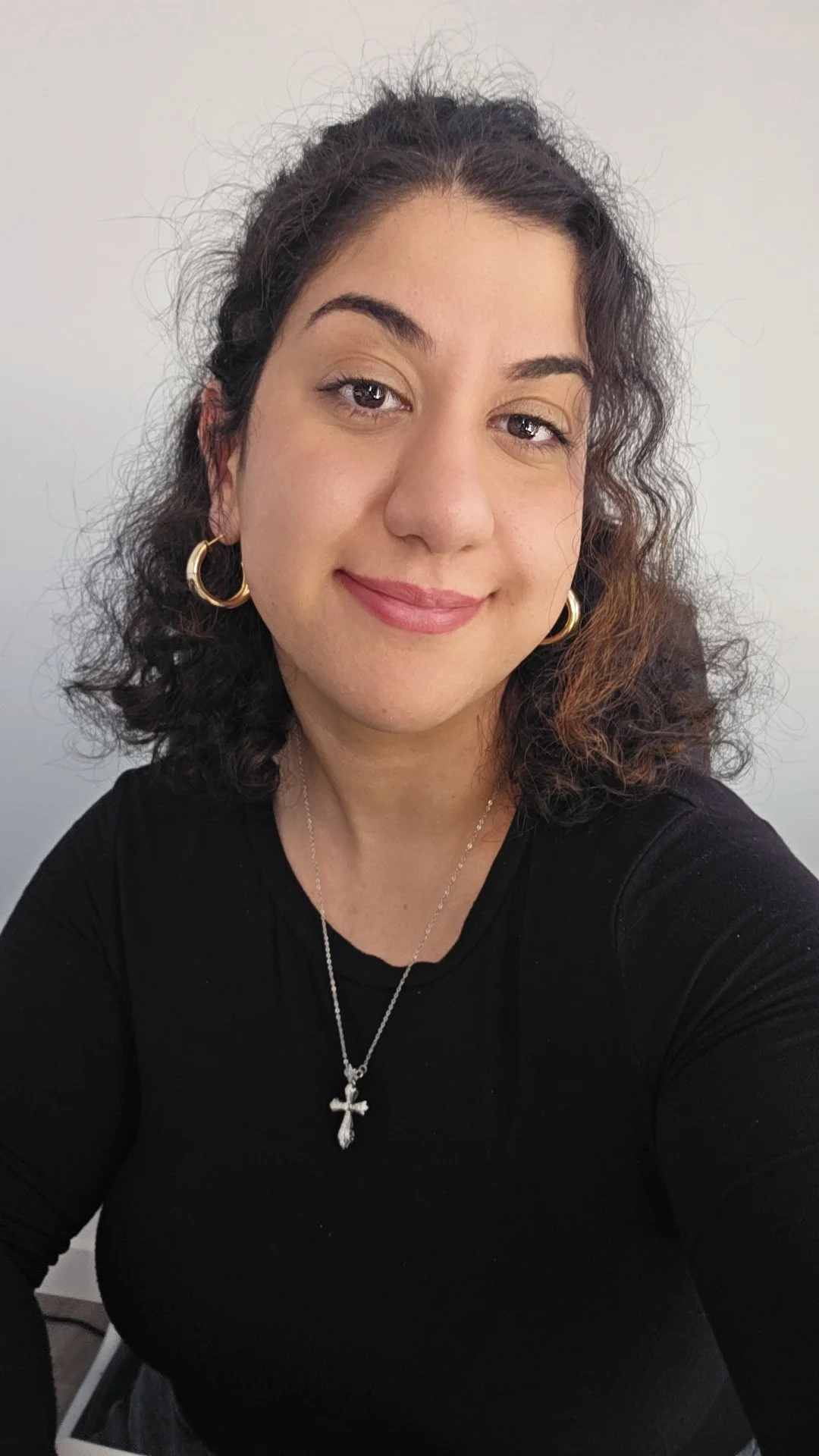 A woman with curly dark hair, wearing a black top, gold hoop earrings, a silver necklace with a cross pendant, and smiling at the camera, in front of a plain light-colored wall.