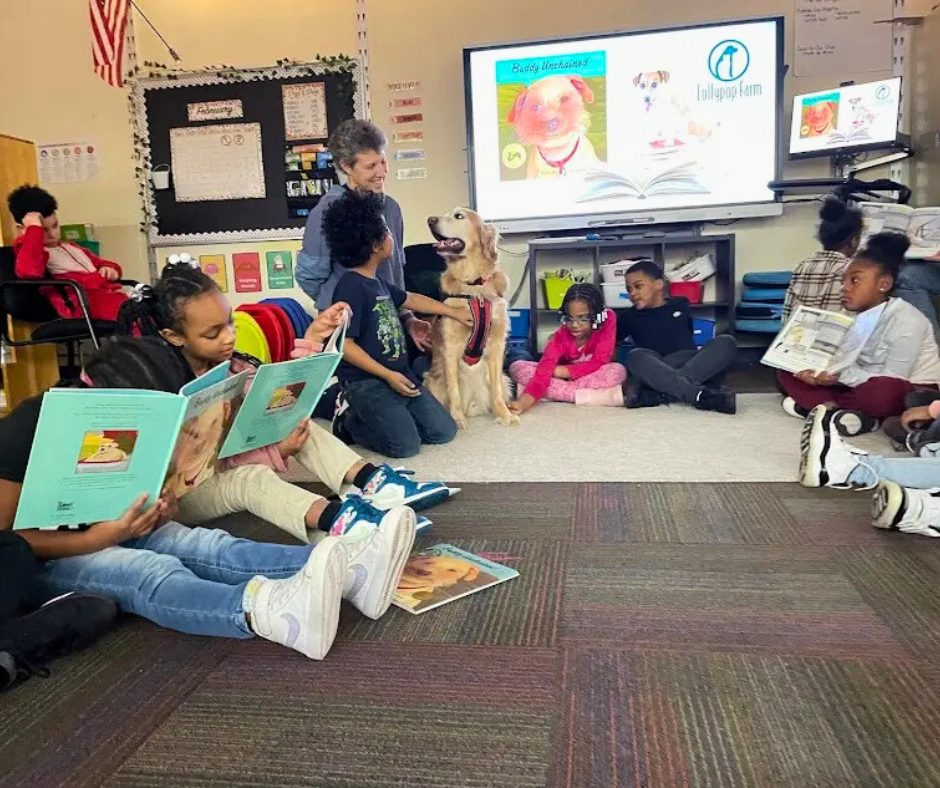 Elementary school classroom with children reading books and a woman and a trained service dog sitting on the floor, a large screen displays a dog illustration and the logo of Lollipoo Farm.
