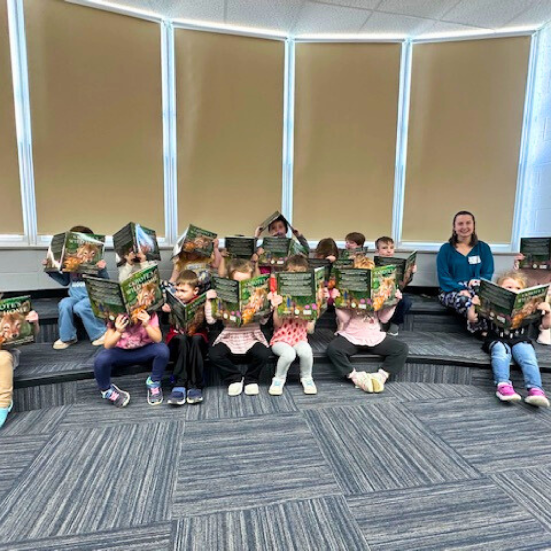 Group of young children sitting on tiered steps, reading animal-themed books, with a smiling adult supervising, in a classroom setting.