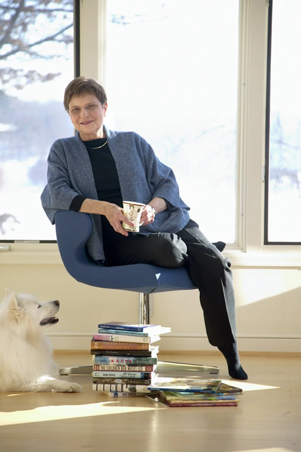 An elderly woman sitting on a blue chair holding a cup, next to a white fluffy dog on the floor, with a stack of books nearby, in a room with large windows and a snowy outdoor scene.
