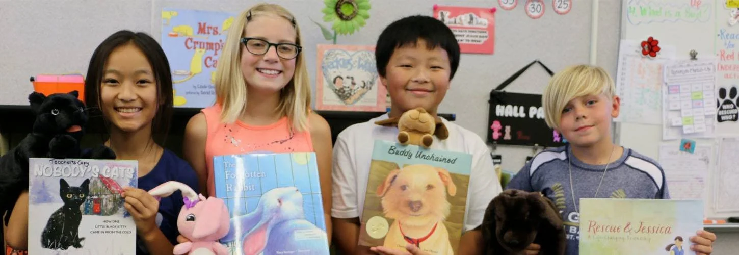 Four children standing in a classroom, each holding a different pet or a children's book related to animals, with colorful posters and papers on the wall behind them.