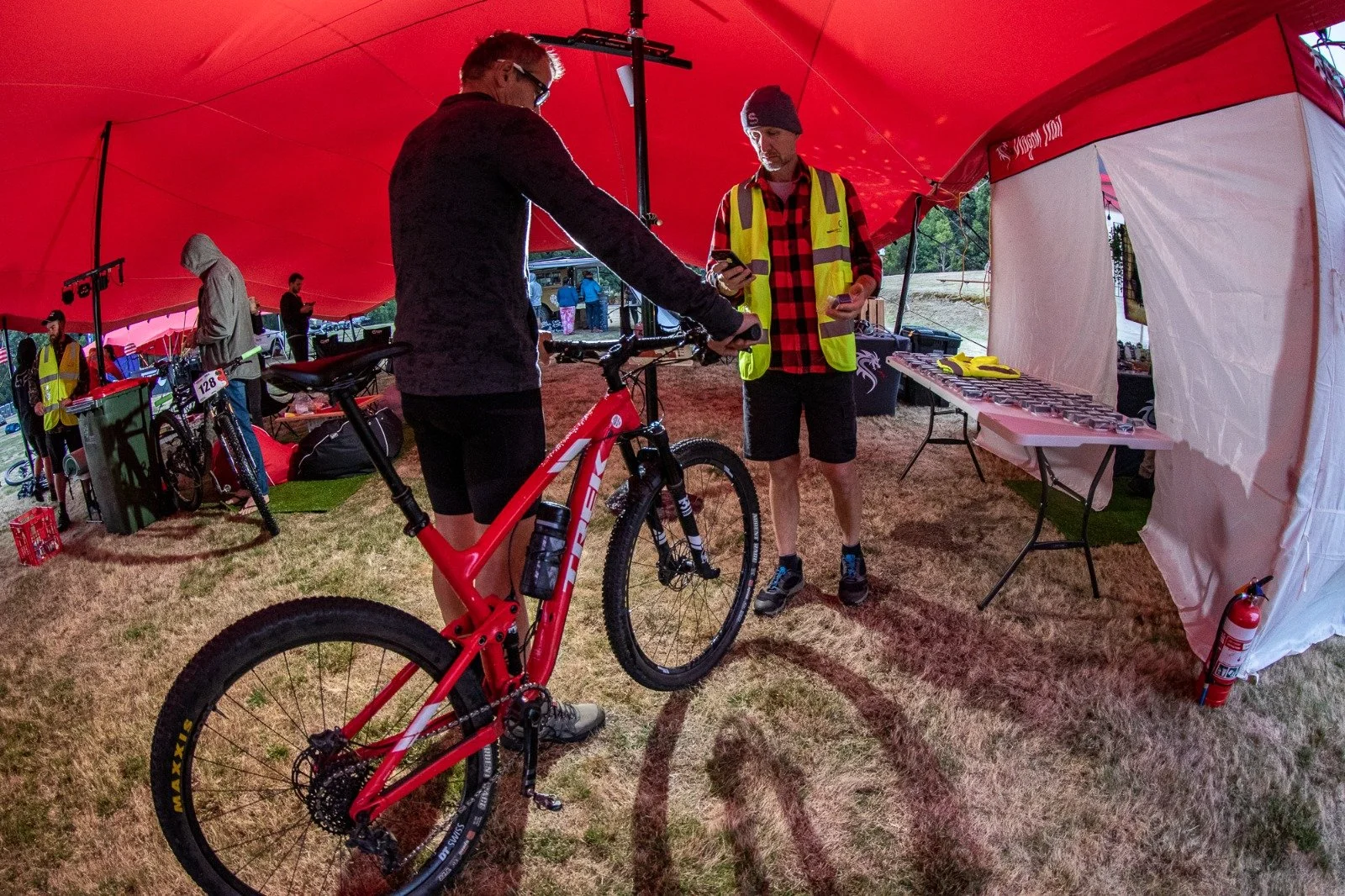 Two men under a red stretch marquee at a cycling event, with one holding a red mountain bike and the other showing a phone, surrounded by other bikes and event supplies.