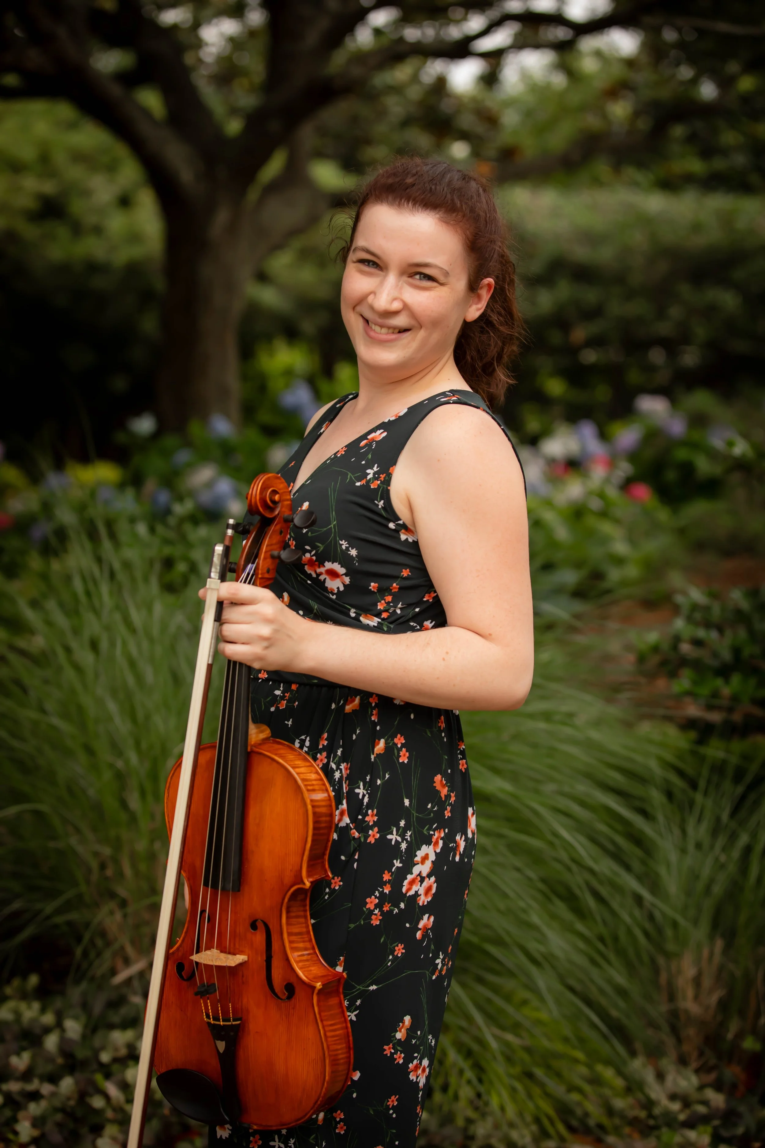 A young woman with curly brown hair, wearing a black floral dress, holding a violin and bow in an outdoor garden setting with green plants and trees.
