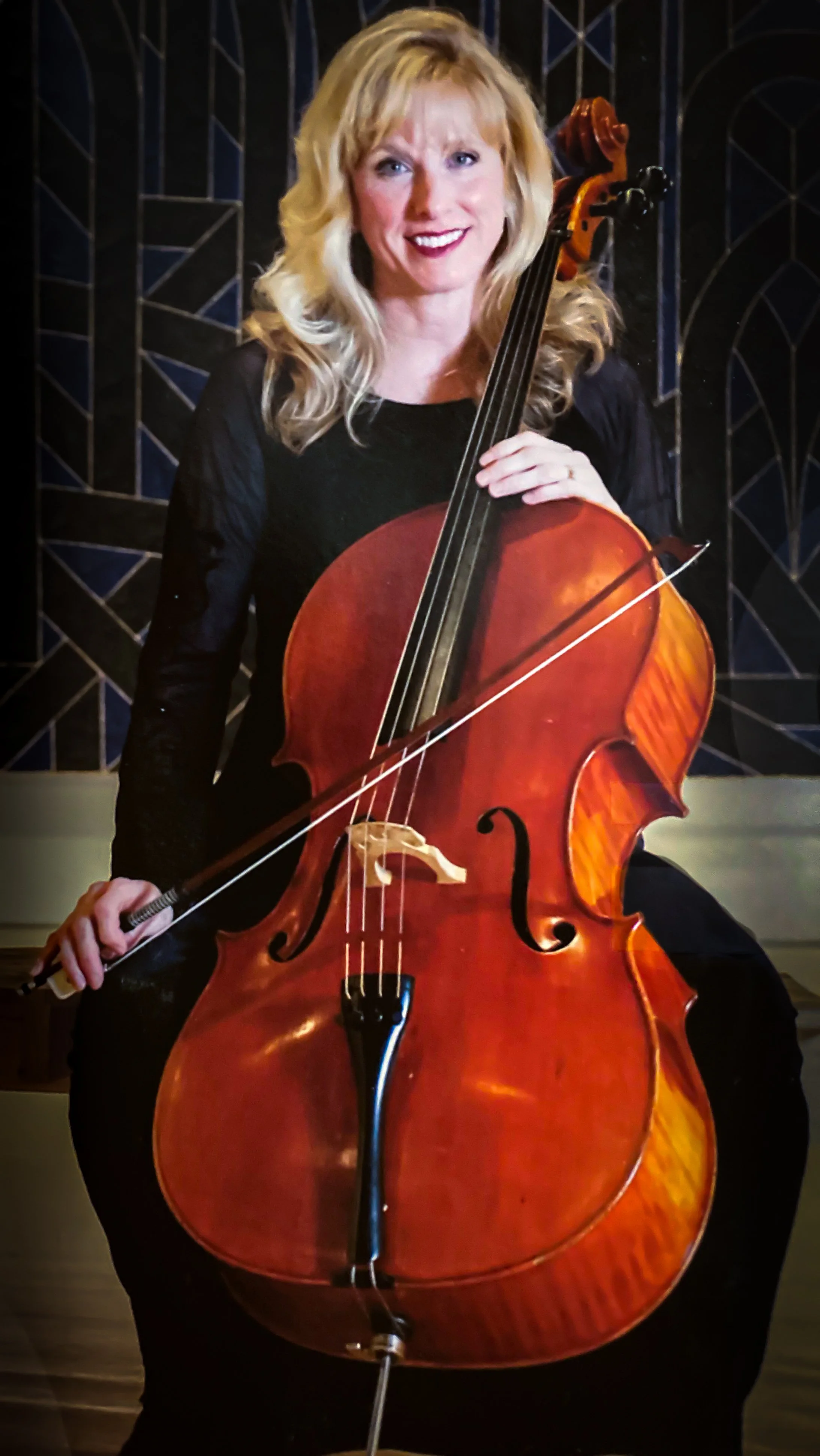 A woman with blonde hair holding a cello and a bow, smiling, in a room with stained glass windows.