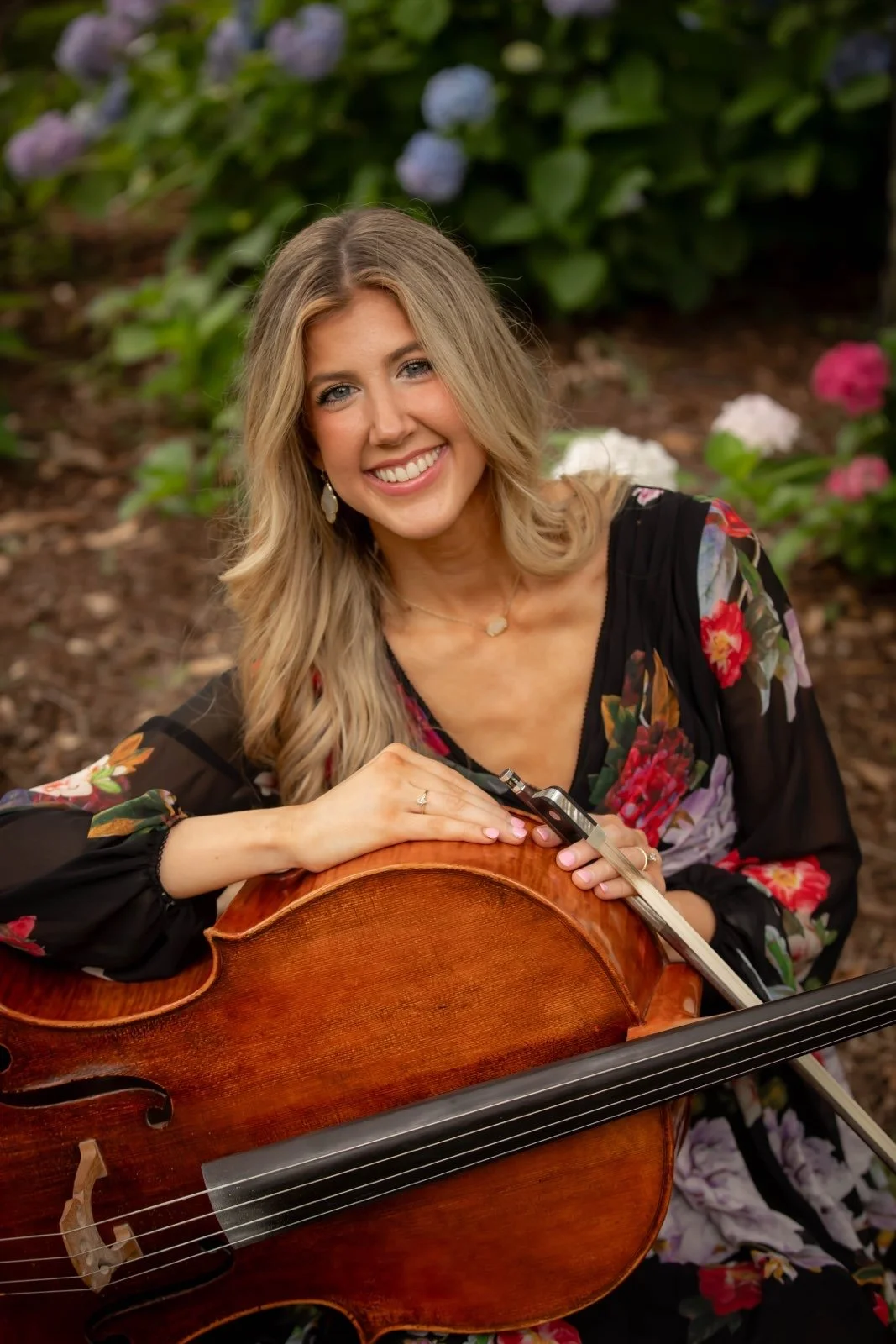 A young woman with blonde hair and a floral dress, smiling and holding a cello in a garden with blooming flowers.