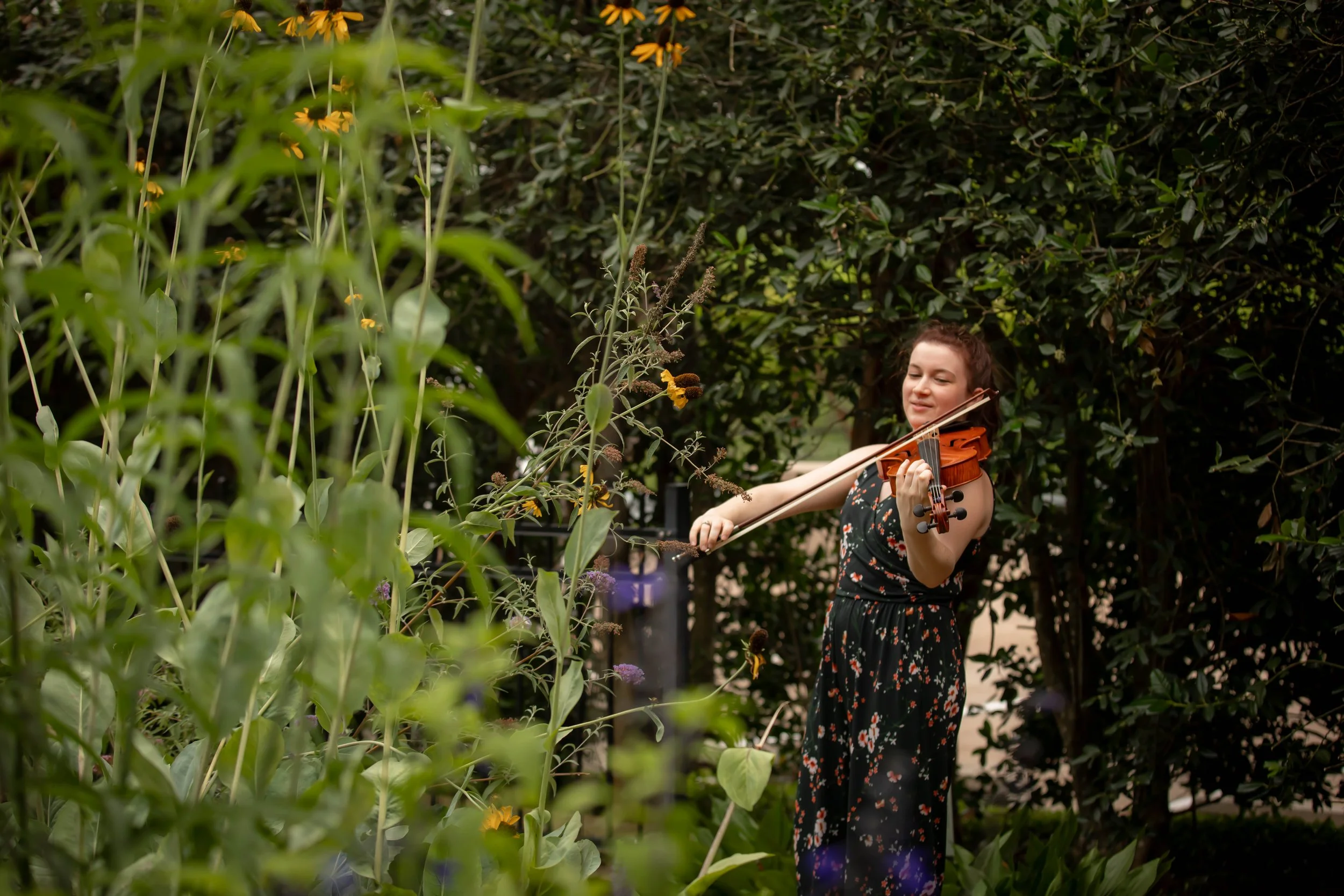 A woman in a dark floral dress playing violin music outdoors in a garden in Mississippi.