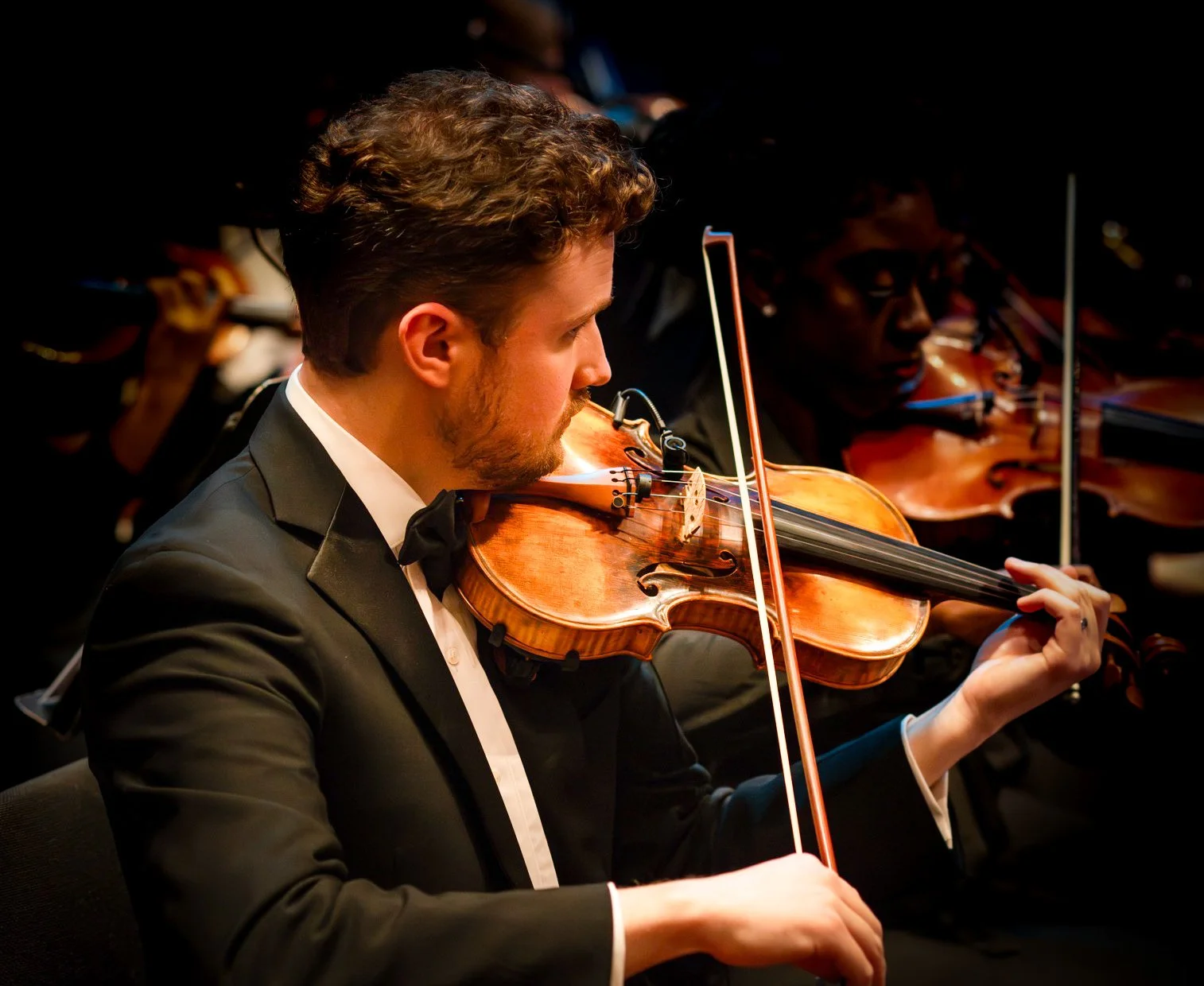A young man in a black tuxedo playing violin in an orchestra concert.