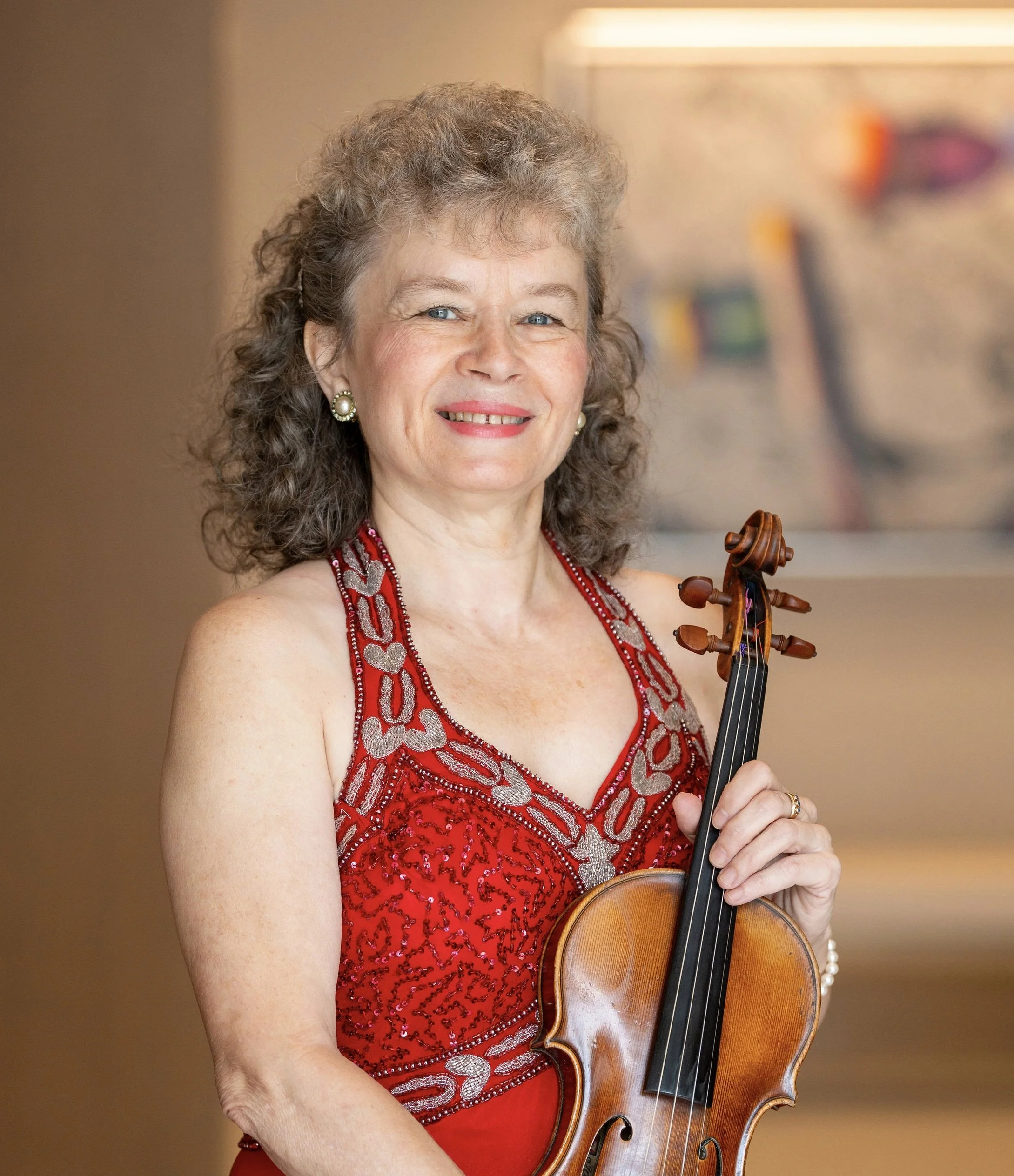 A smiling woman with curly hair wearing a red evening gown holding a violin.
