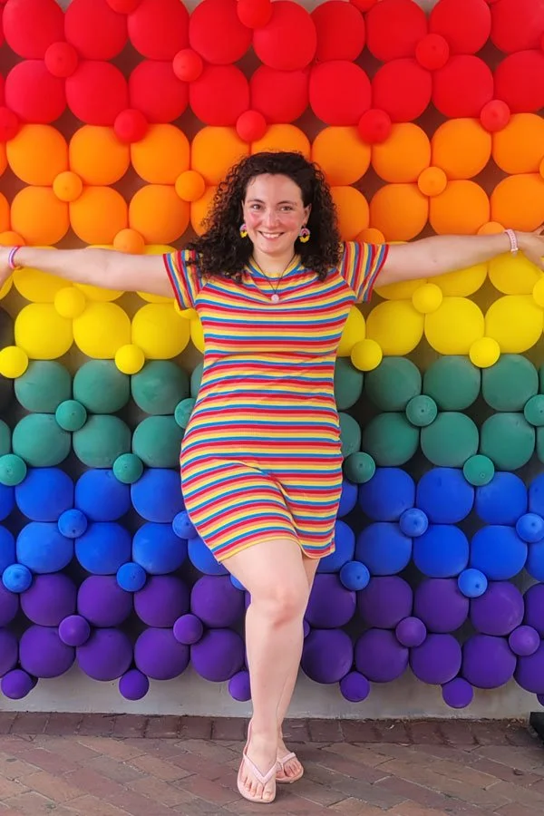 Portrait of artist, Anastasia Longoria. A young person with shoulder-length curly black hair. She smiles, wearing a mid-thigh rainbow striped dress, standing arms outstretched in front of a rainbow balloon wall.