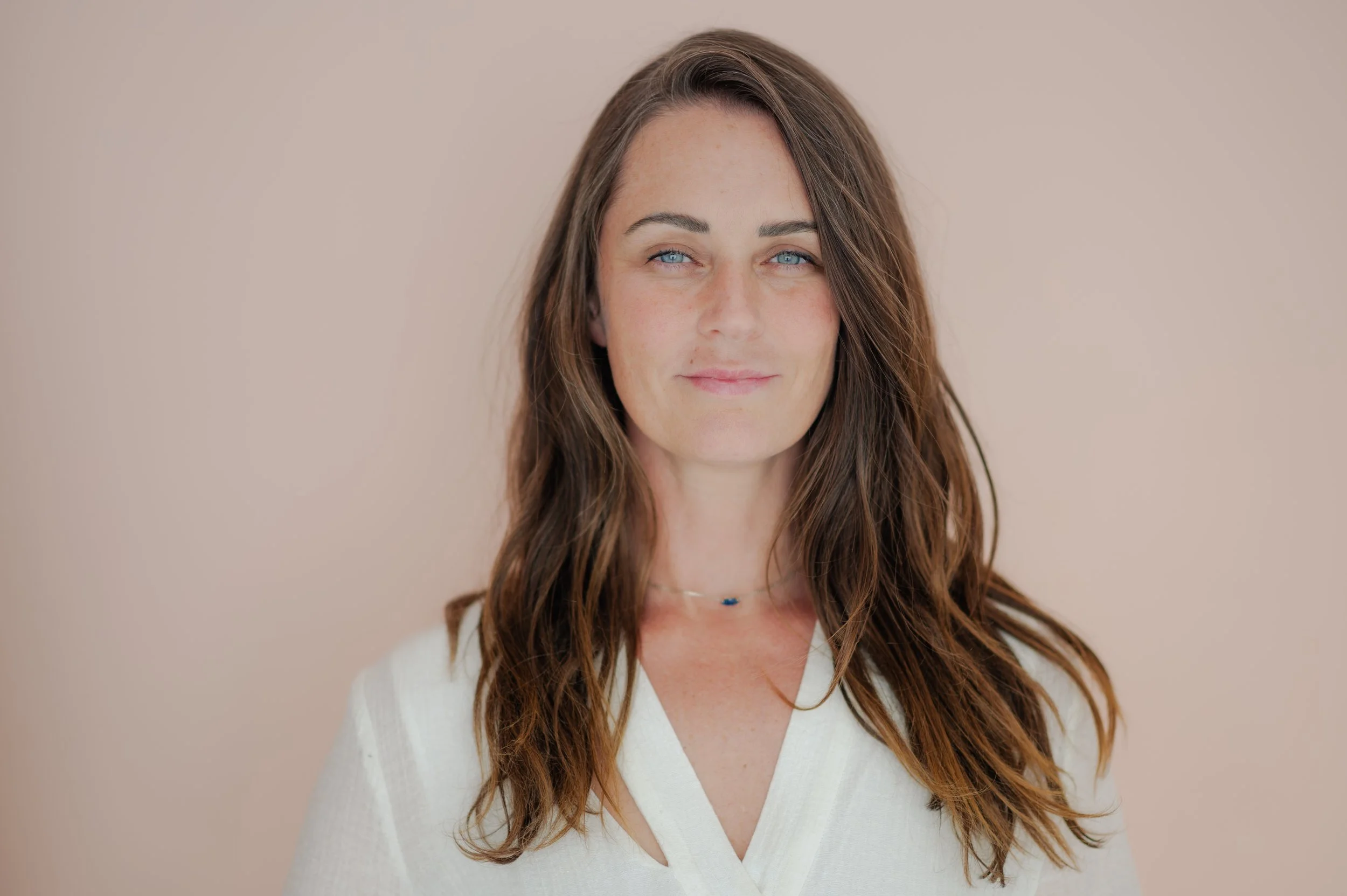 A woman with long, wavy brown hair and blue eyes, wearing a white V-neck top and a delicate necklace, standing against a neutral beige background.