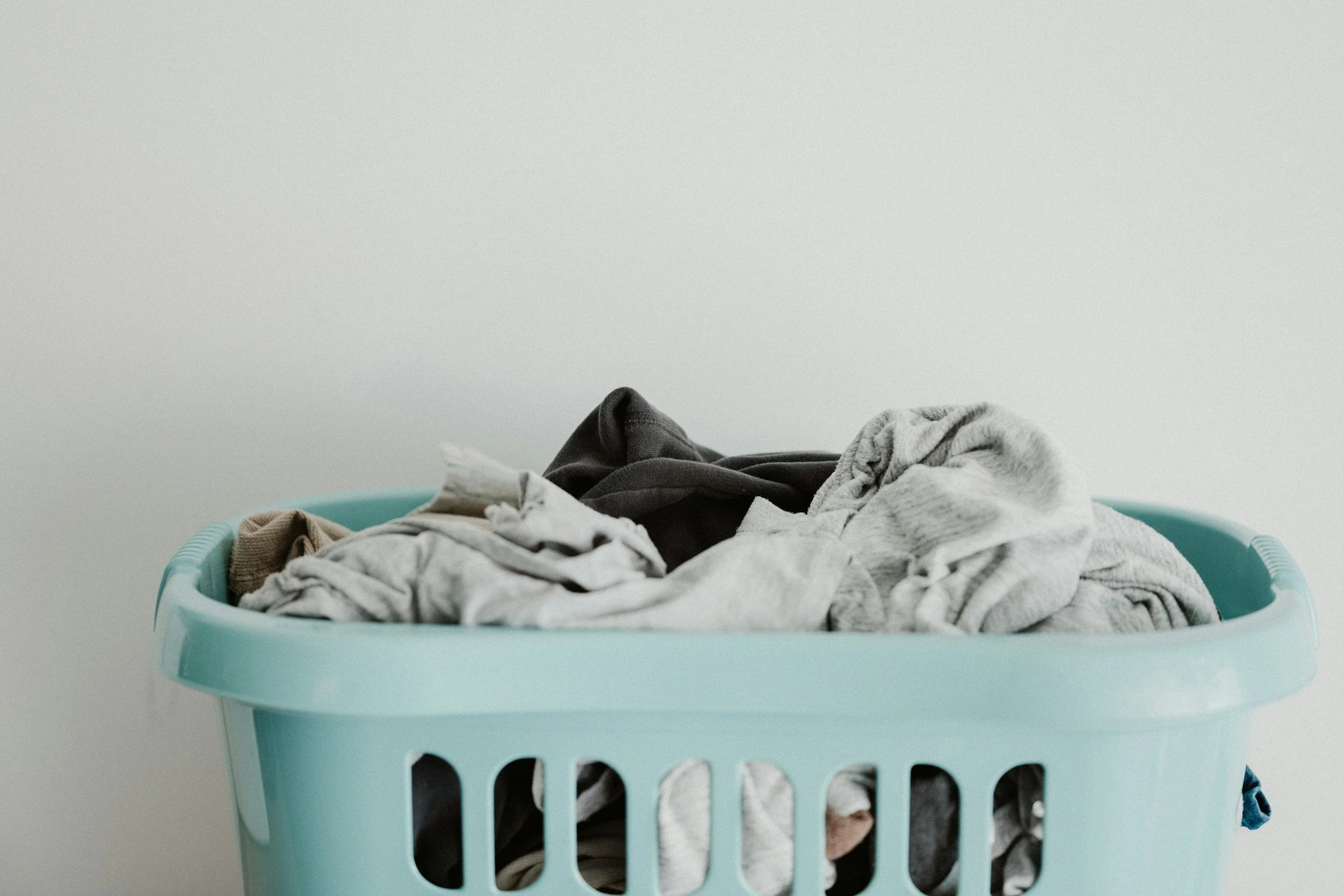 Light blue laundry basket filled with mixed, wrinkled clothes against a plain light-colored wall.