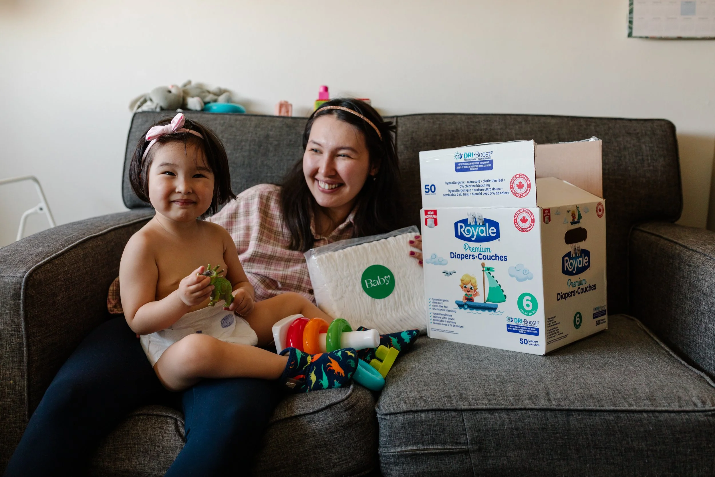 A mom and her baby sit on a couch. The baby is playing with some toys and the mom is holding a pack of Royale Premium Diapers she received from Baby Love.