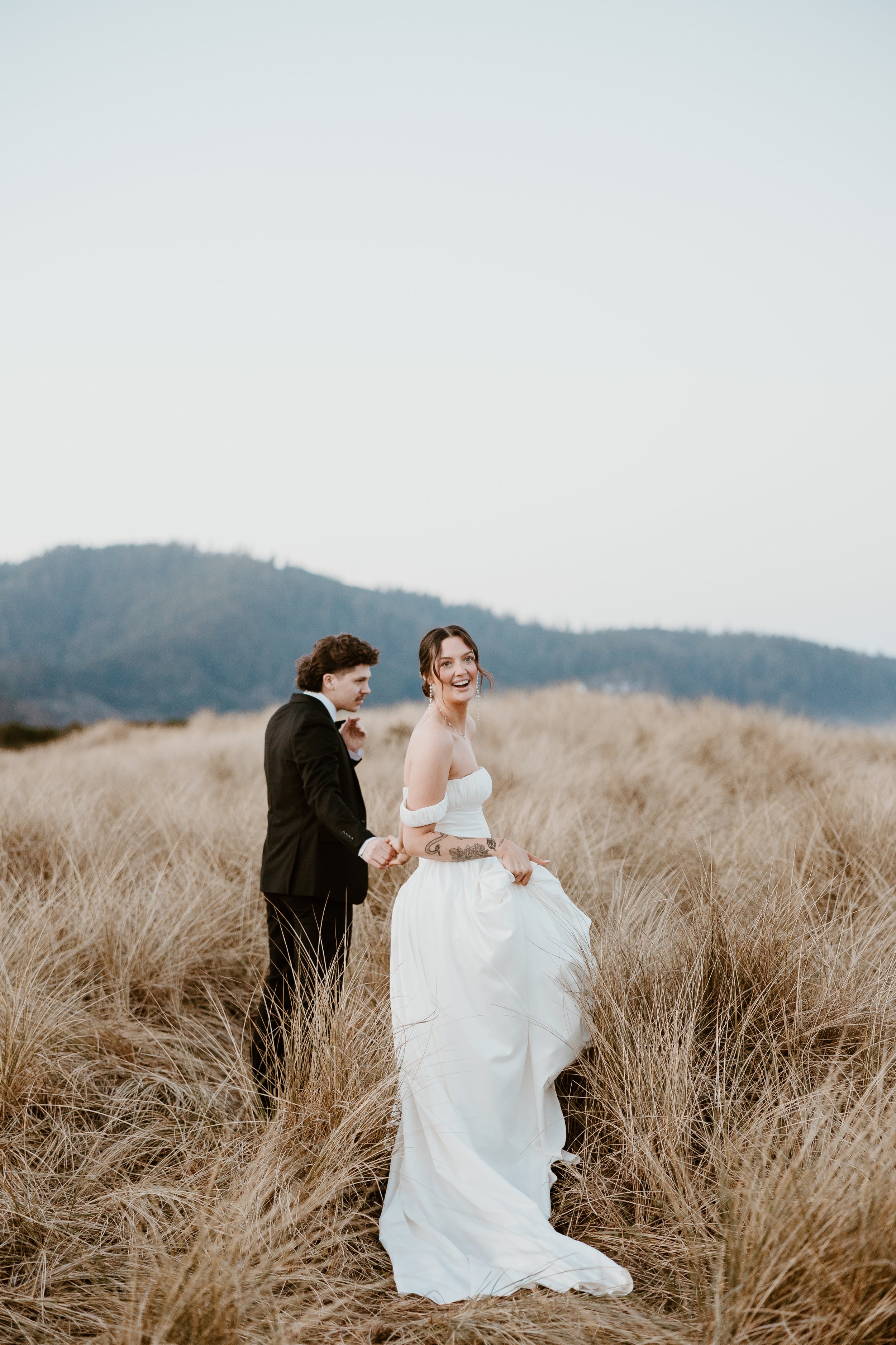 A woman in a white wedding dress and a man in a black tuxedo standing in a grassy field, with mountains in the background and a clear sky above.