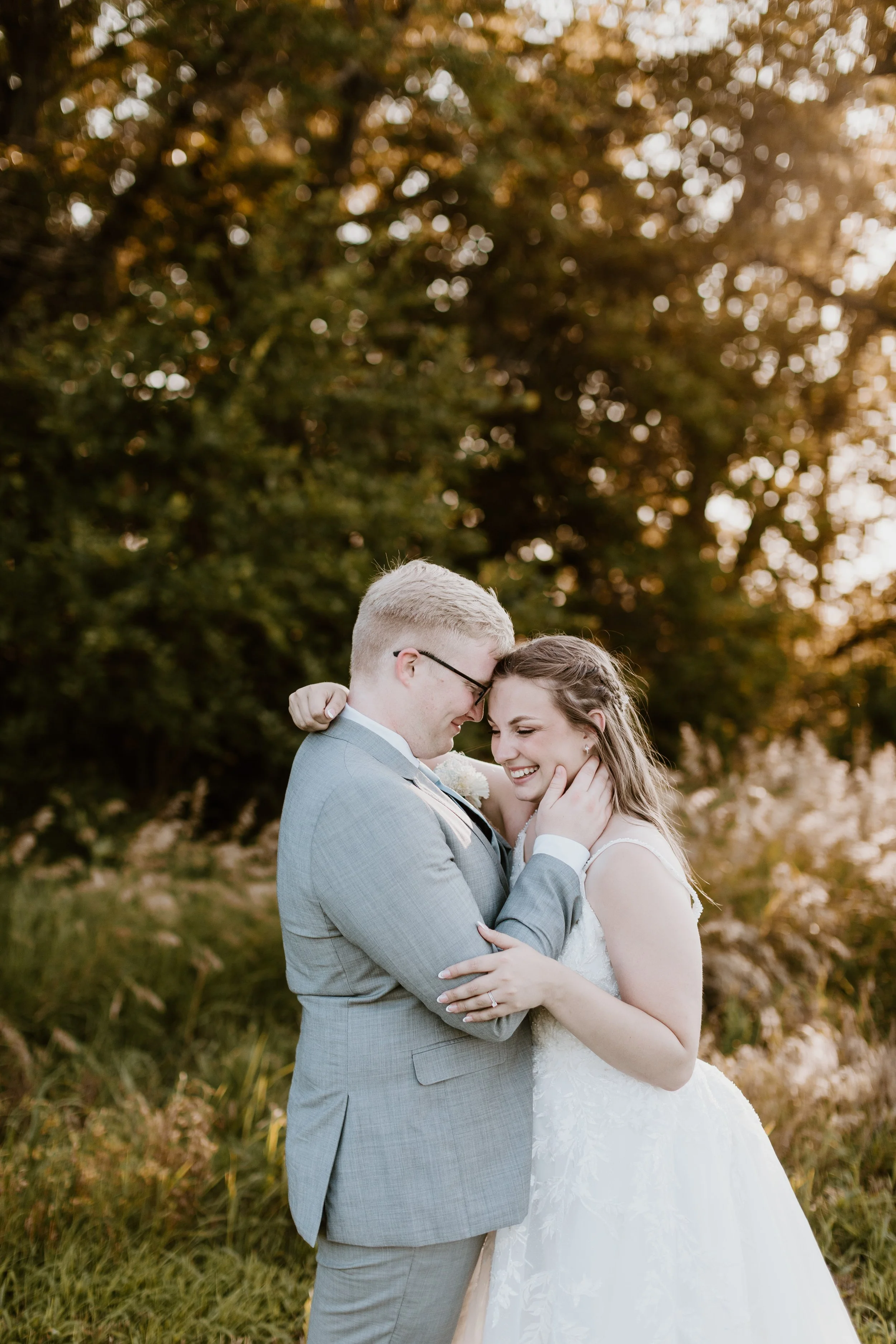 A couple, dressed in wedding attire, embraces and smiles outdoors during sunset, with a background of trees and golden light.