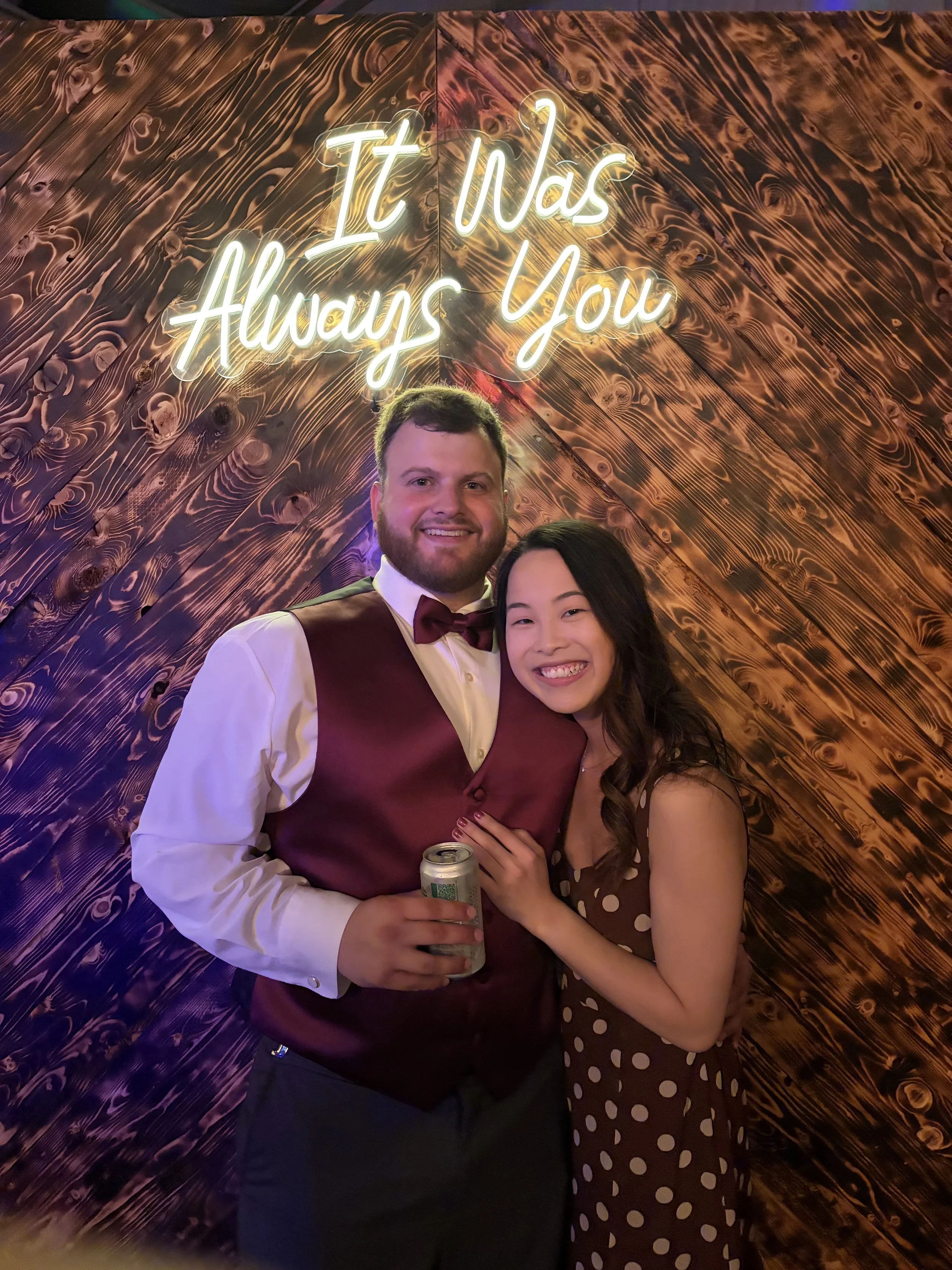 A smiling man and woman embrace in front of a wooden wall with neon sign reading 'It Was Always You'. The man is wearing a white shirt, burgundy vest, and bow tie, holding a can. The woman is in a brown polka dot dress.