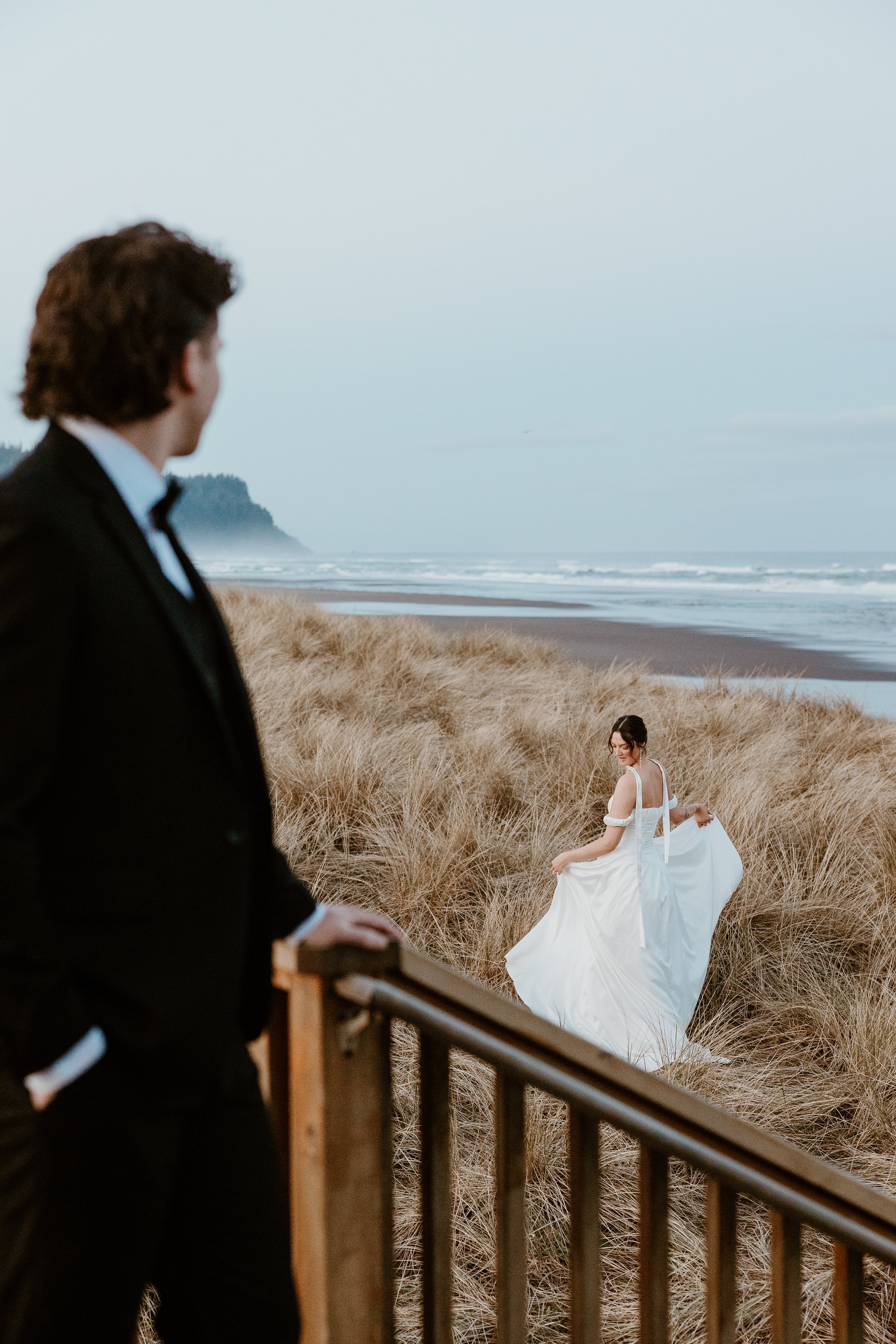 A man in a tuxedo looking at a woman in a white wedding dress on a beach with tall grasses and the ocean in the background.