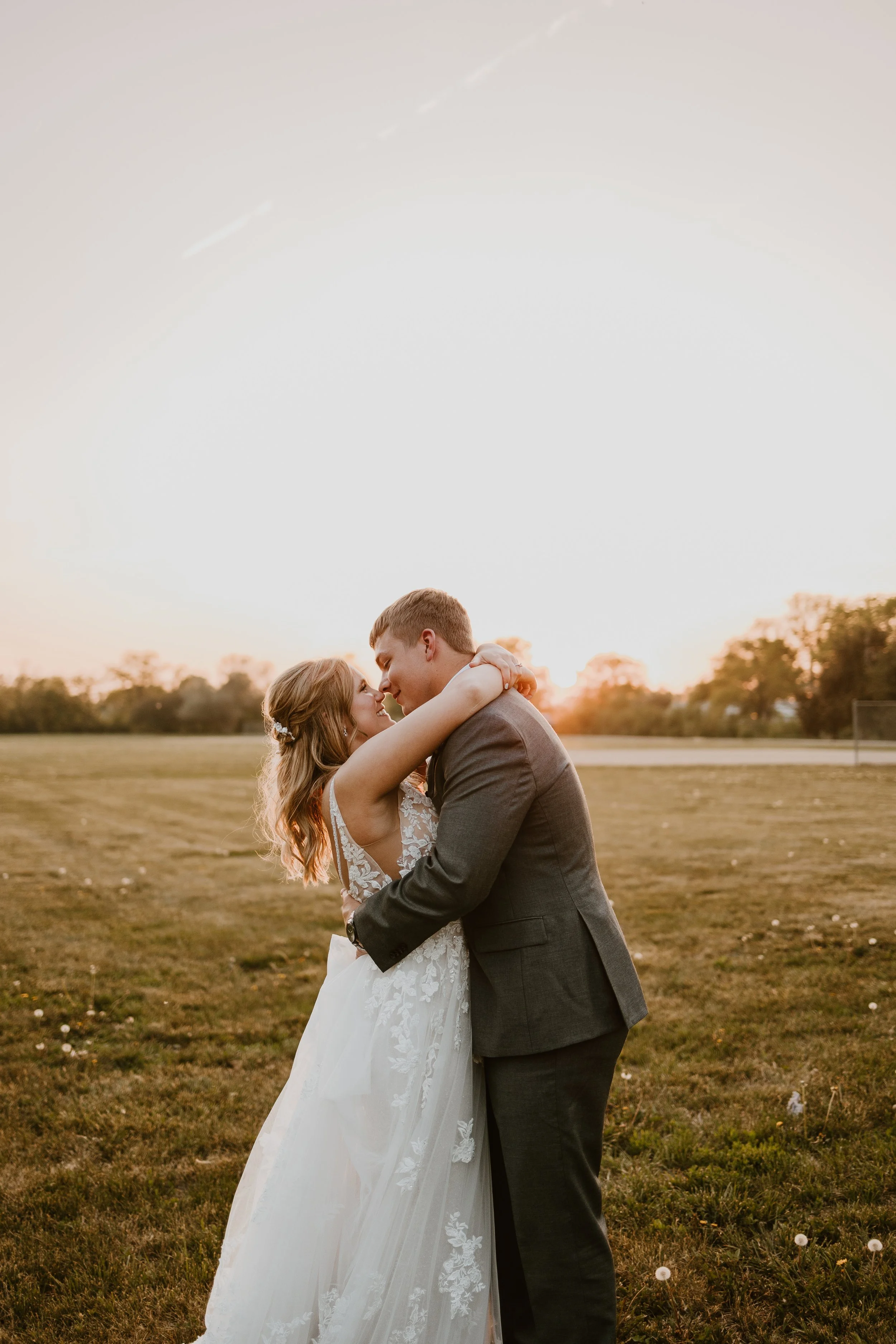 A couple in wedding attire embracing outdoors at sunset, with trees and a field in the background.