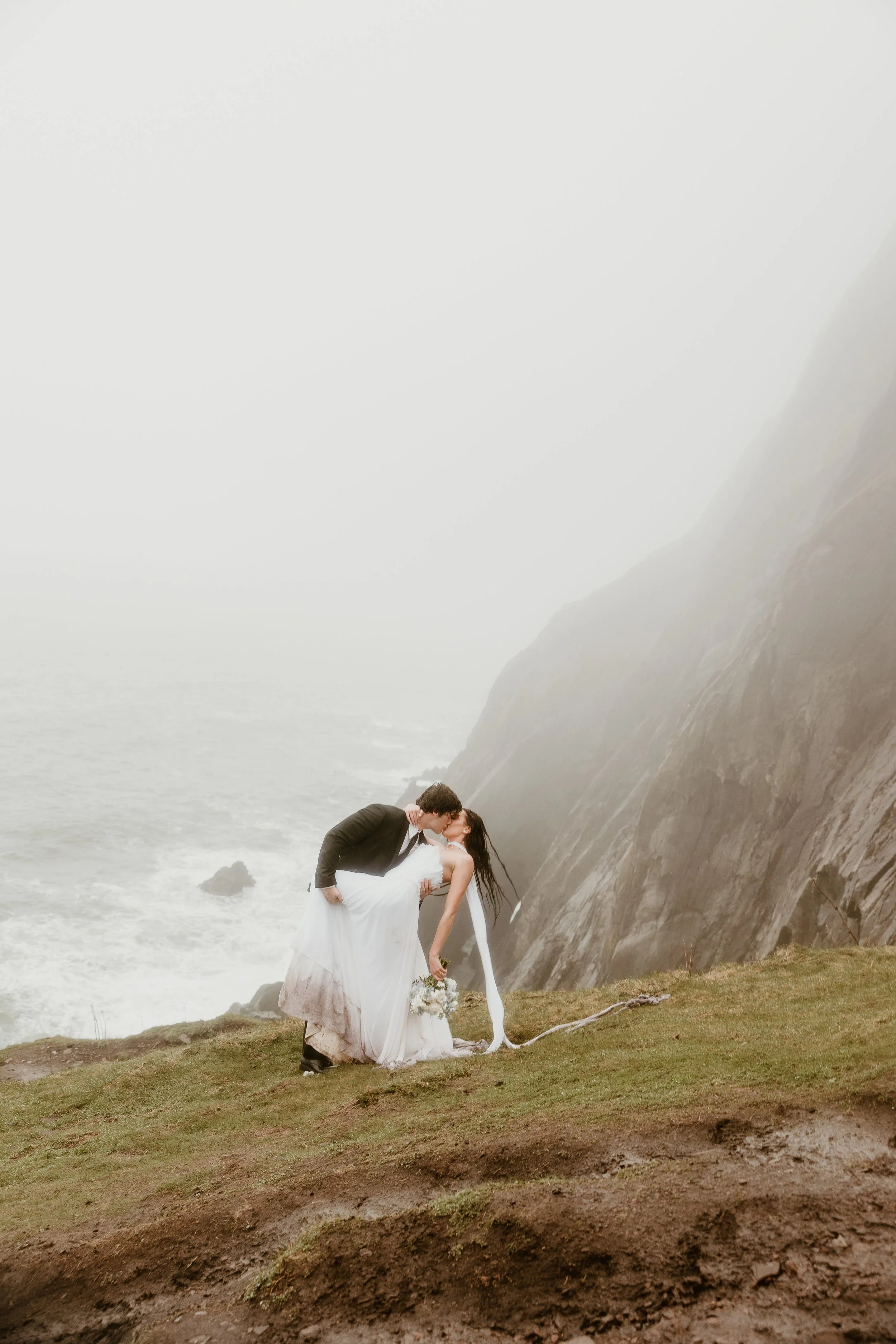 A couple in wedding attire sharing a kiss on a grassy cliff overlooking the ocean with foggy weather.