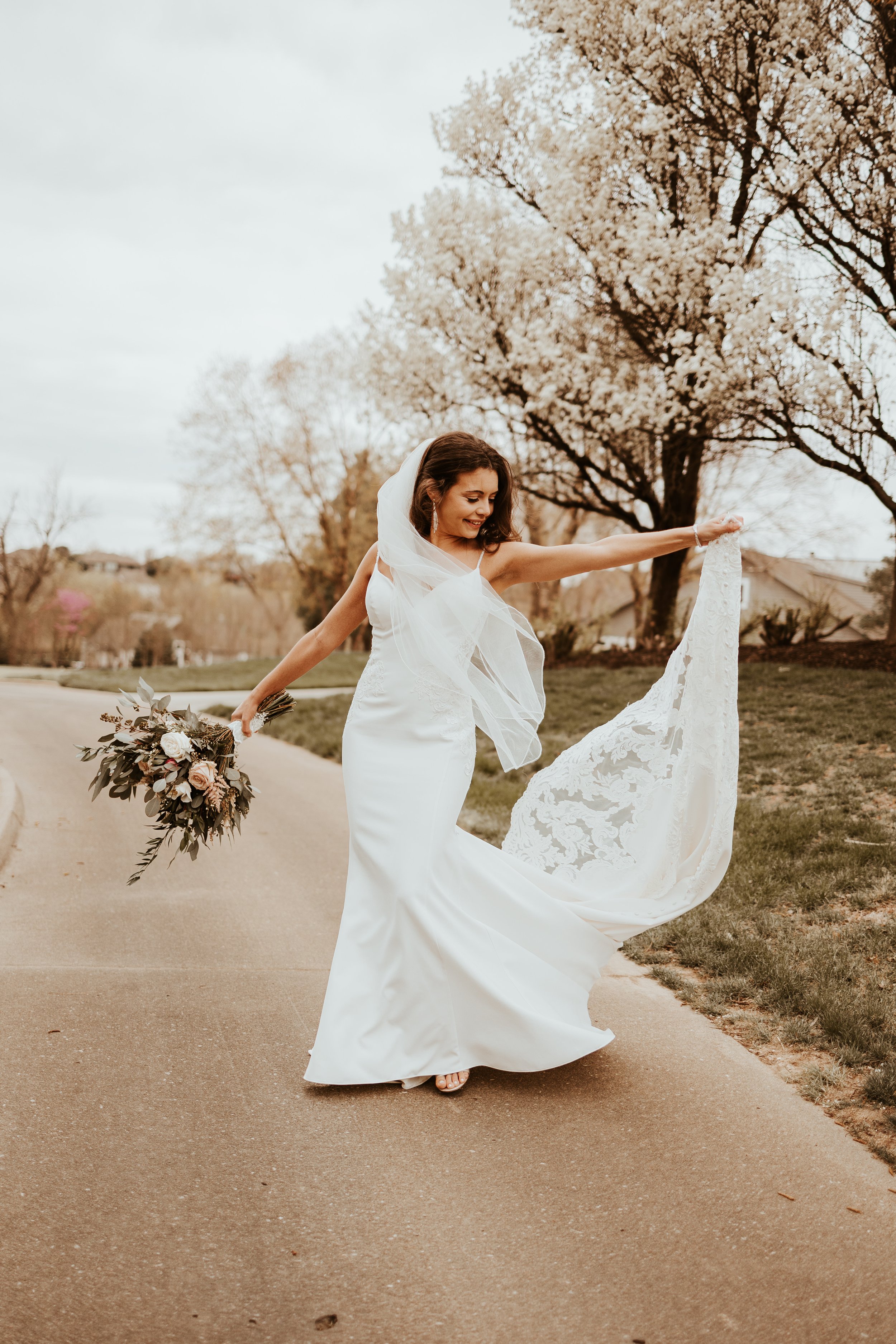 A bride in a white wedding dress holding her train and a bouquet while standing on a paved path with blooming trees in the background.
