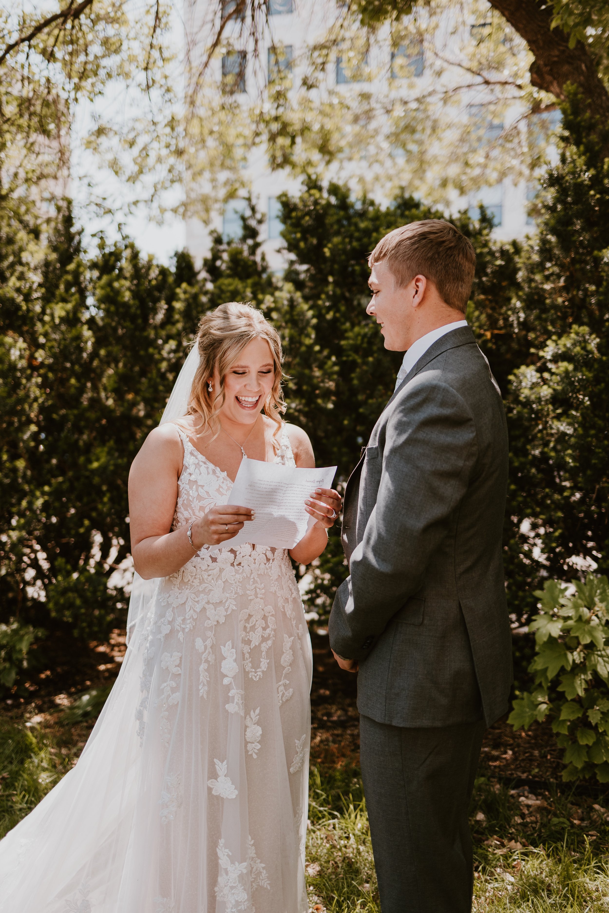 A bride and groom at an outdoor wedding ceremony, the bride reading vows from a paper, surrounded by trees and greenery.
