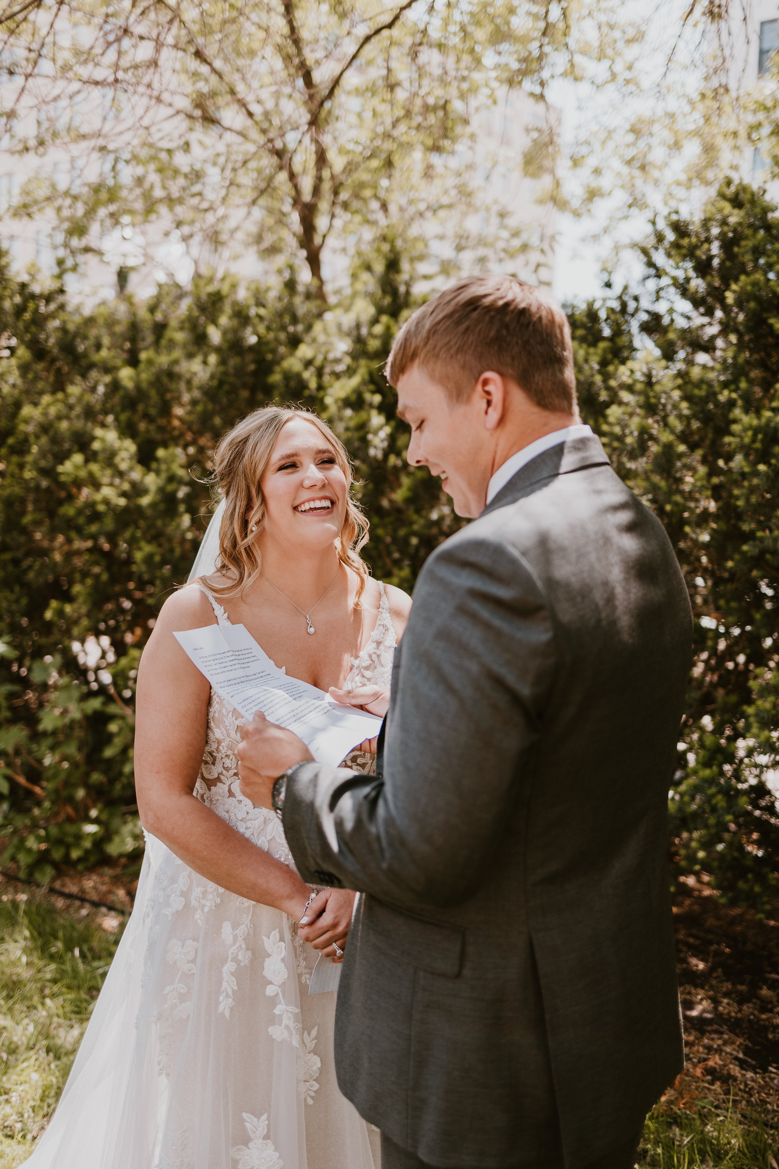 A bride and groom during their wedding vows outdoors, with the groom reading from a paper and the bride smiling.