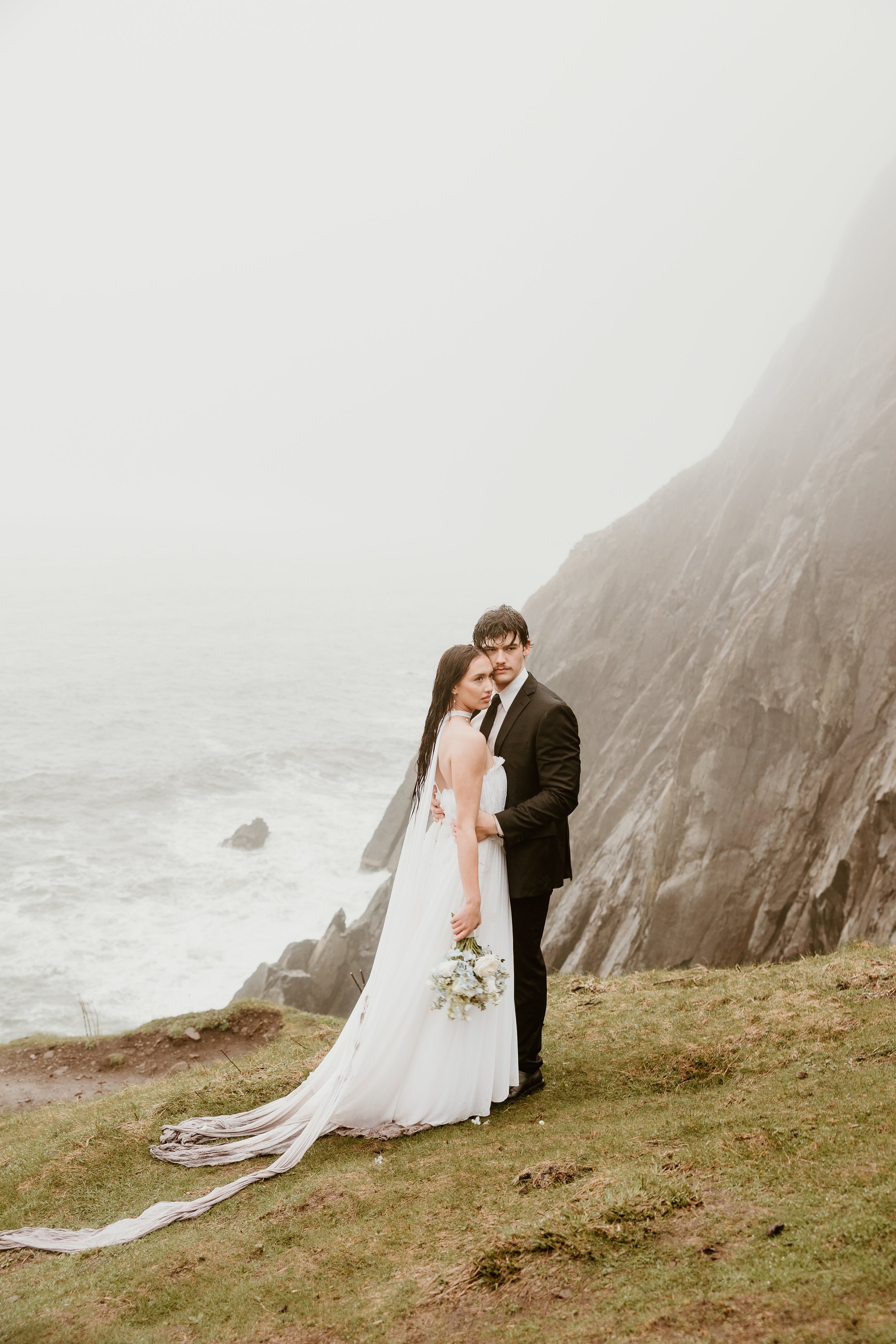 A bride and groom standing close together on a grassy cliffside with a foggy ocean background, the bride holding a bouquet of flowers and wearing a flowing white wedding dress, the groom in a black suit and tie.