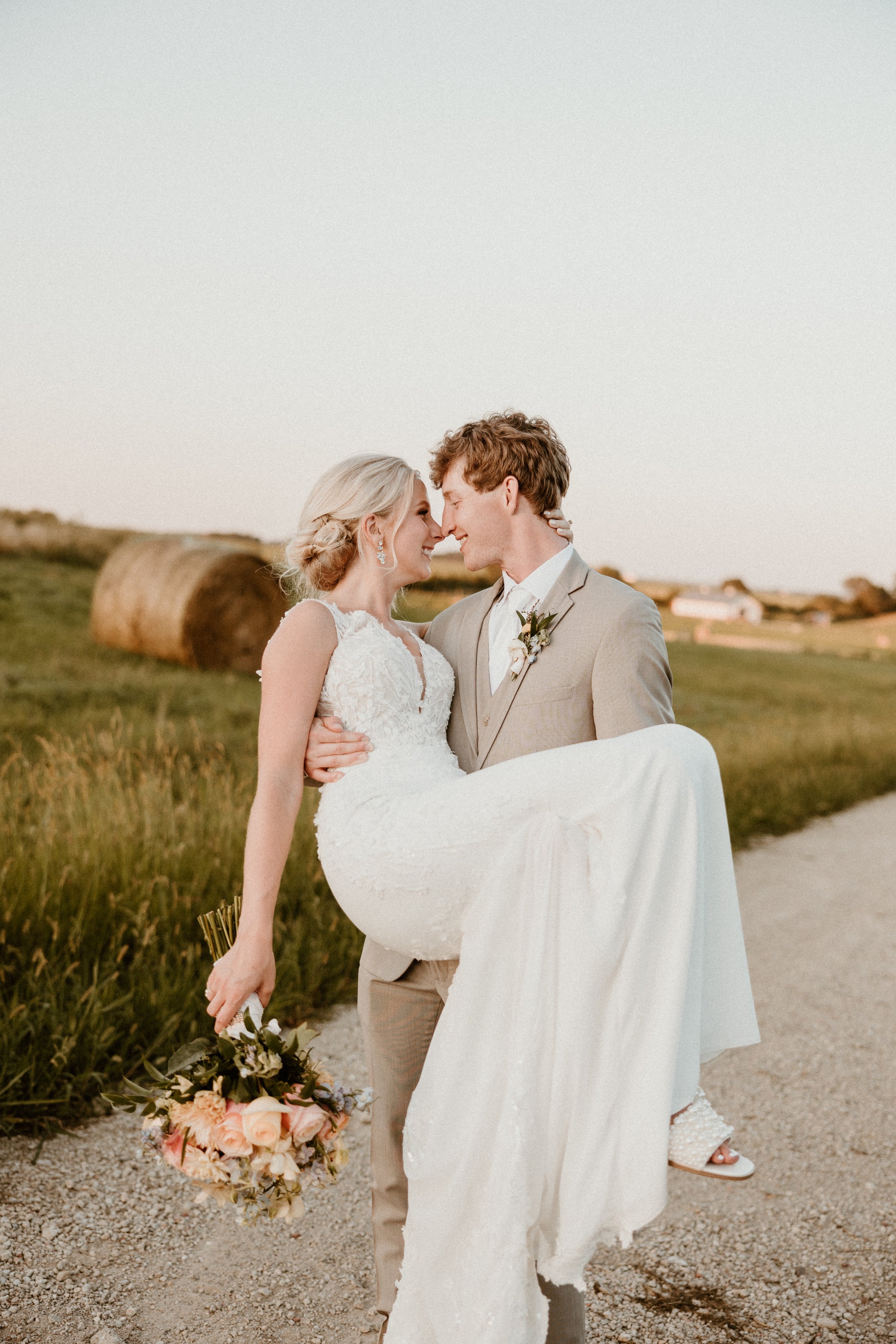 A bride in a white lace wedding dress being carried by a groom in a beige suit on a rural dirt road at sunset, with hay bales and fields in the background.