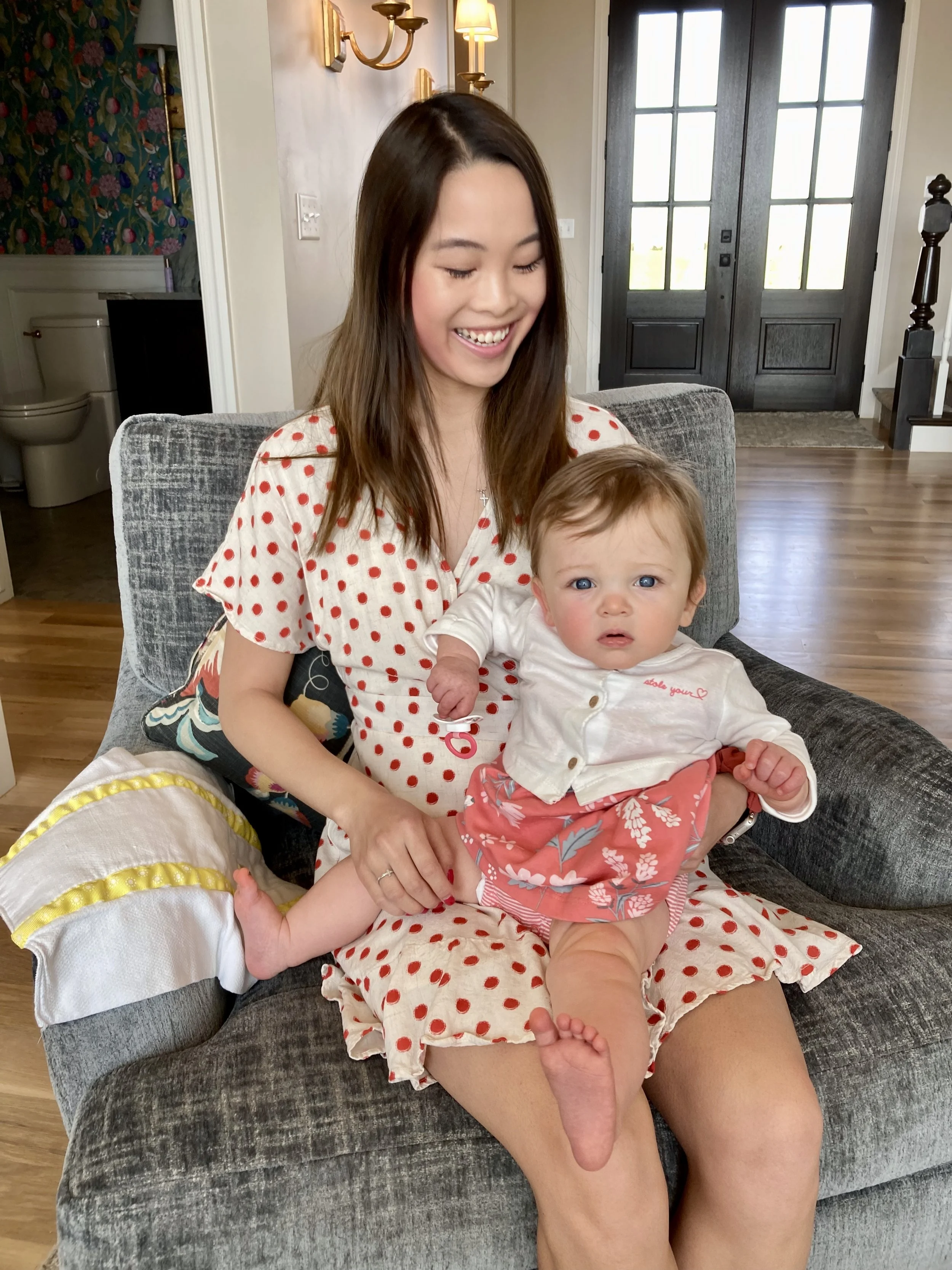 A young woman with long brown hair, wearing a cream-colored dress with red polka dots, sitting on a gray couch with a small baby girl. The woman is smiling while the baby, who has light brown hair and blue eyes, appears curious and is wearing a white