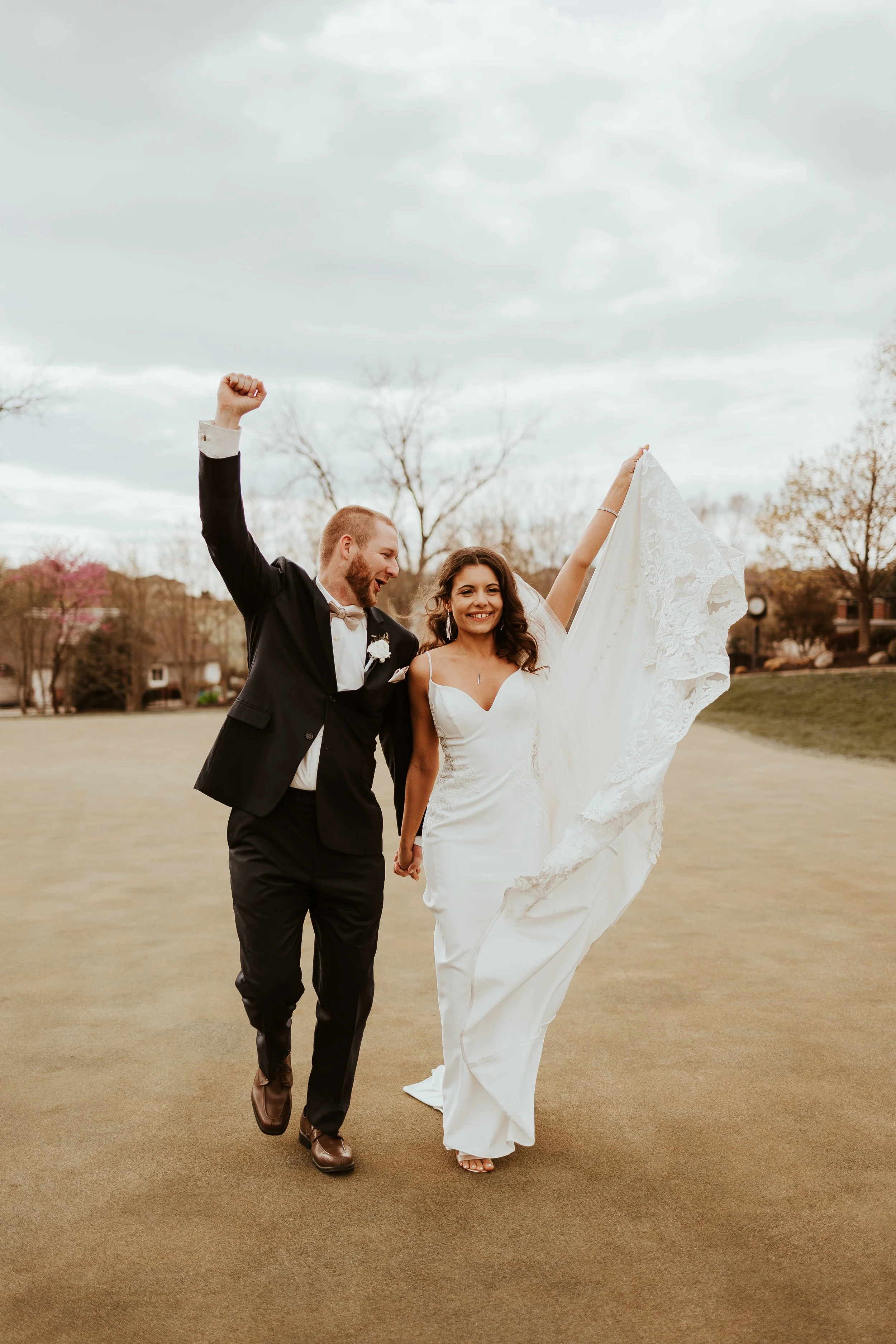 A newlywed couple celebrating outdoors on their wedding day, holding hands and smiling, with the bride lifting her wedding dress.