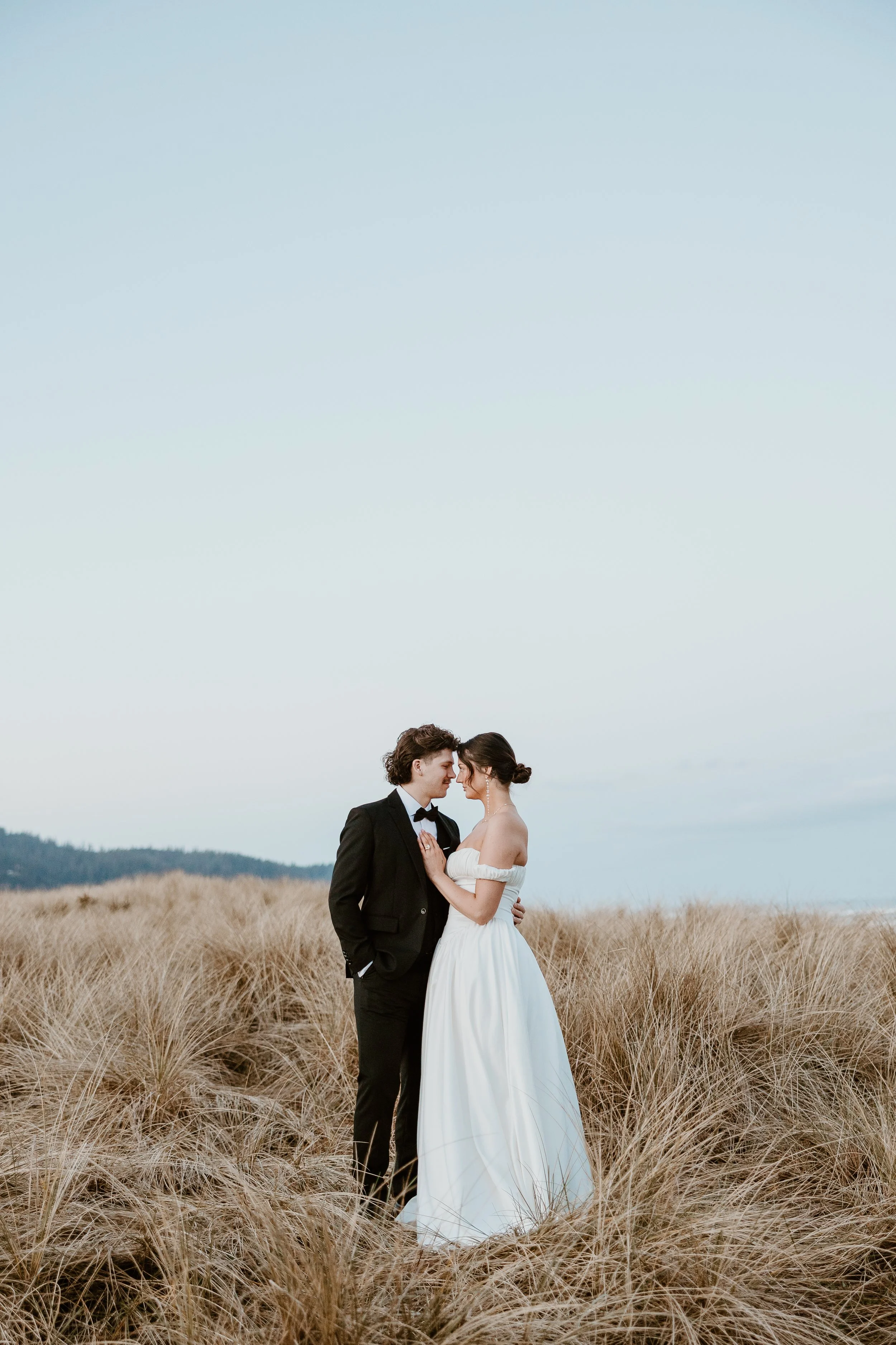 A bride and groom standing close together in a field of tall, dry grass, with a clear sky overhead, during their wedding photoshoot.