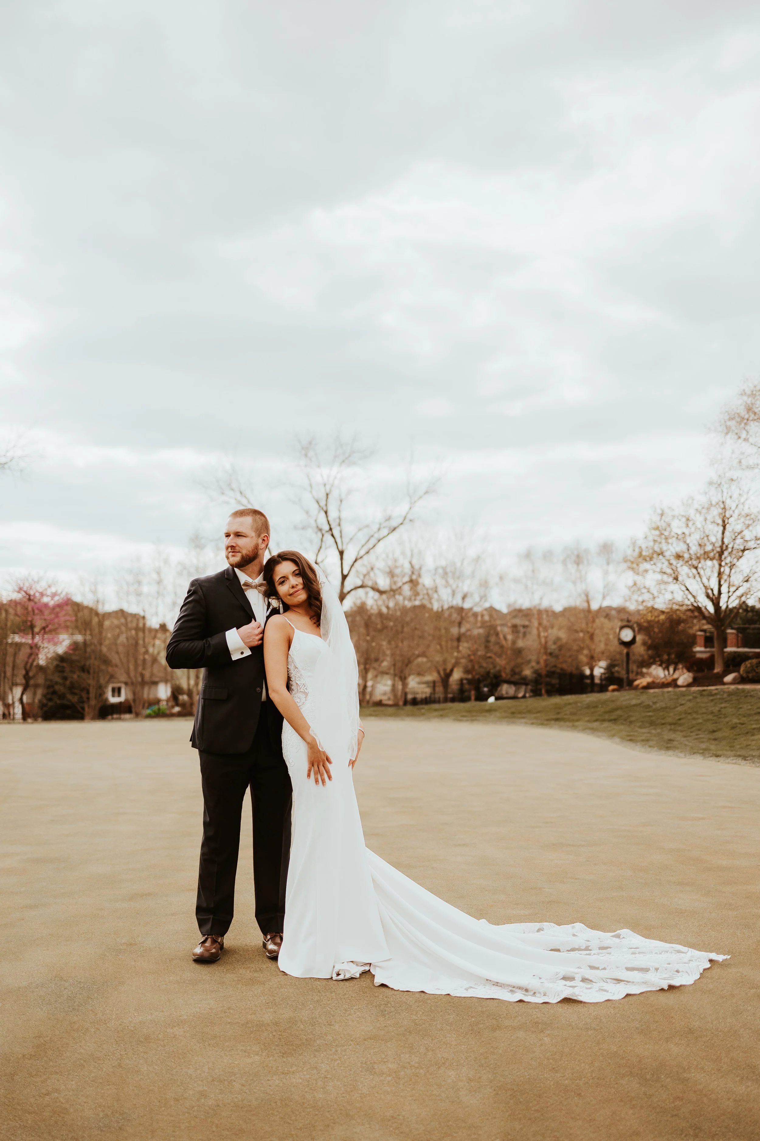 A bride and groom standing outdoors on a cloudy day, with the bride in a white wedding gown and veil, and the groom in a black tuxedo, on a golf course or park.