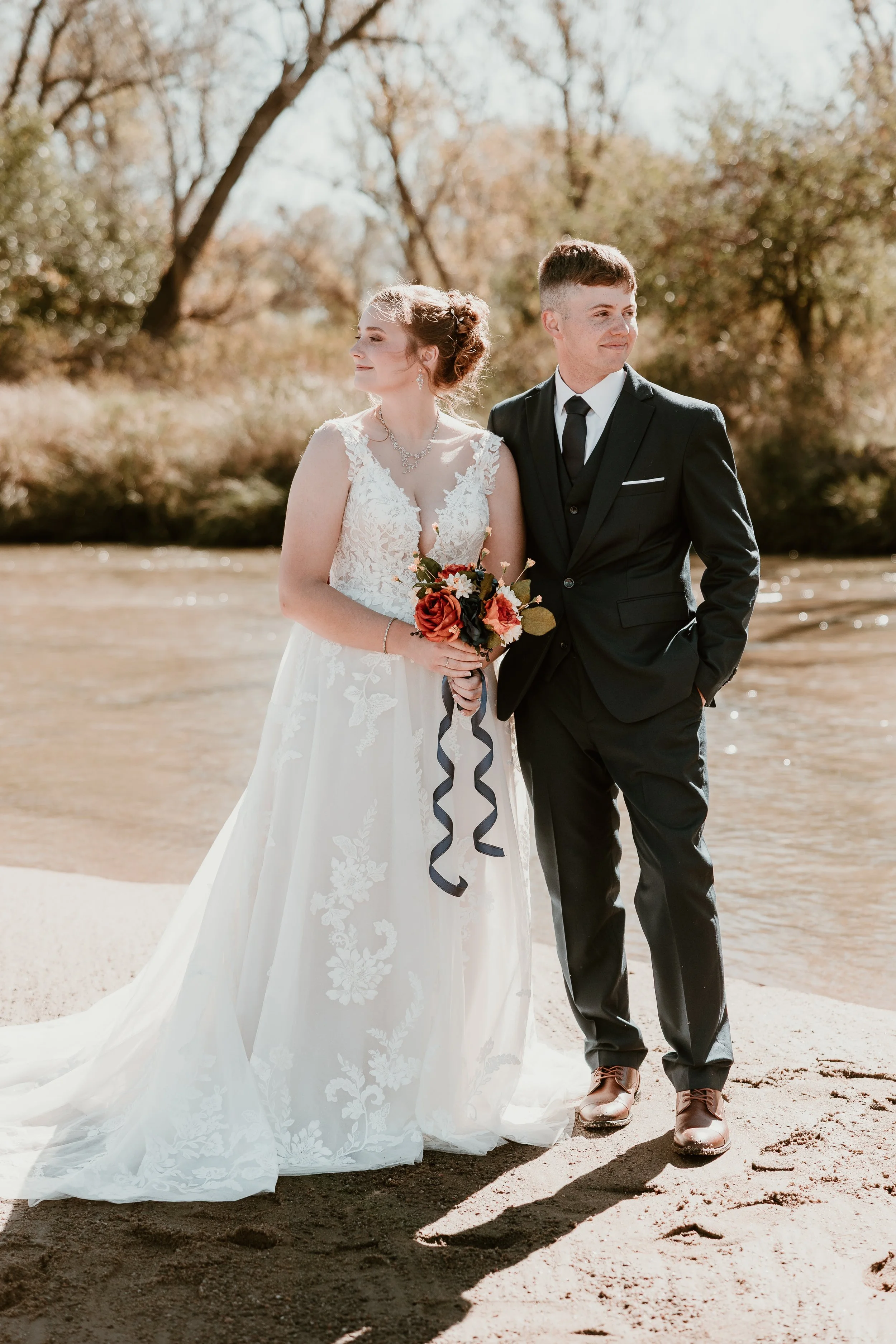 A bride in a white lace wedding dress holding a bouquet standing next to a groom in a black suit by a river with autumn trees in the background.