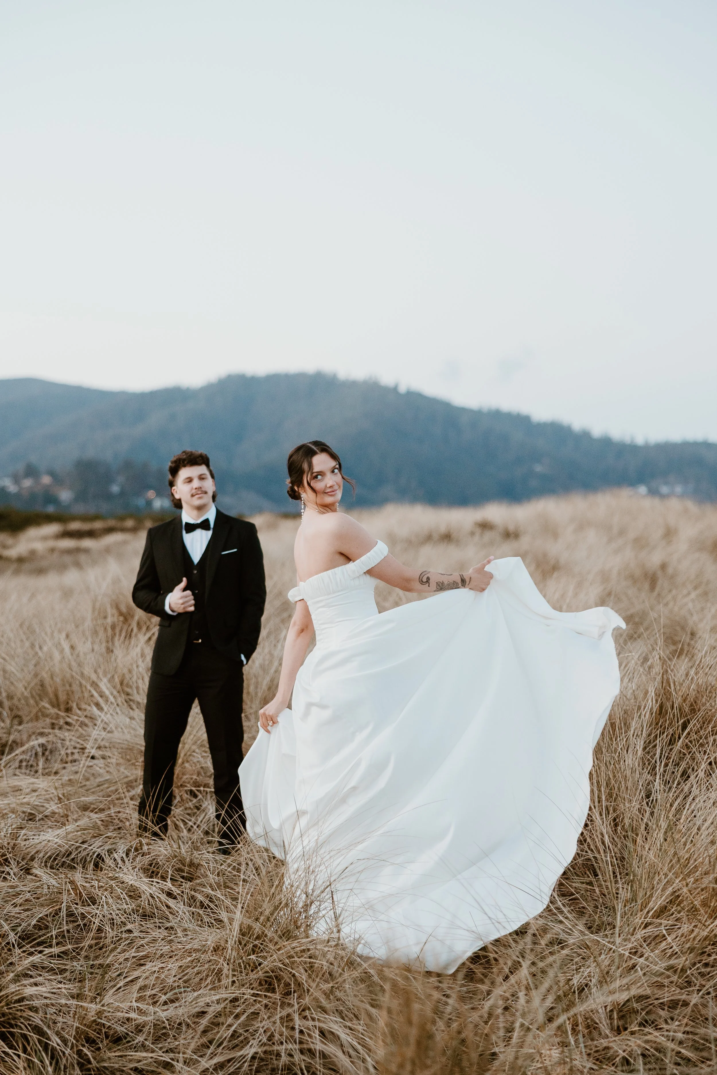 A bride in a white wedding dress holding up her dress while standing in a field of tall grass, with a groom in a black tuxedo in the background during the daytime.
