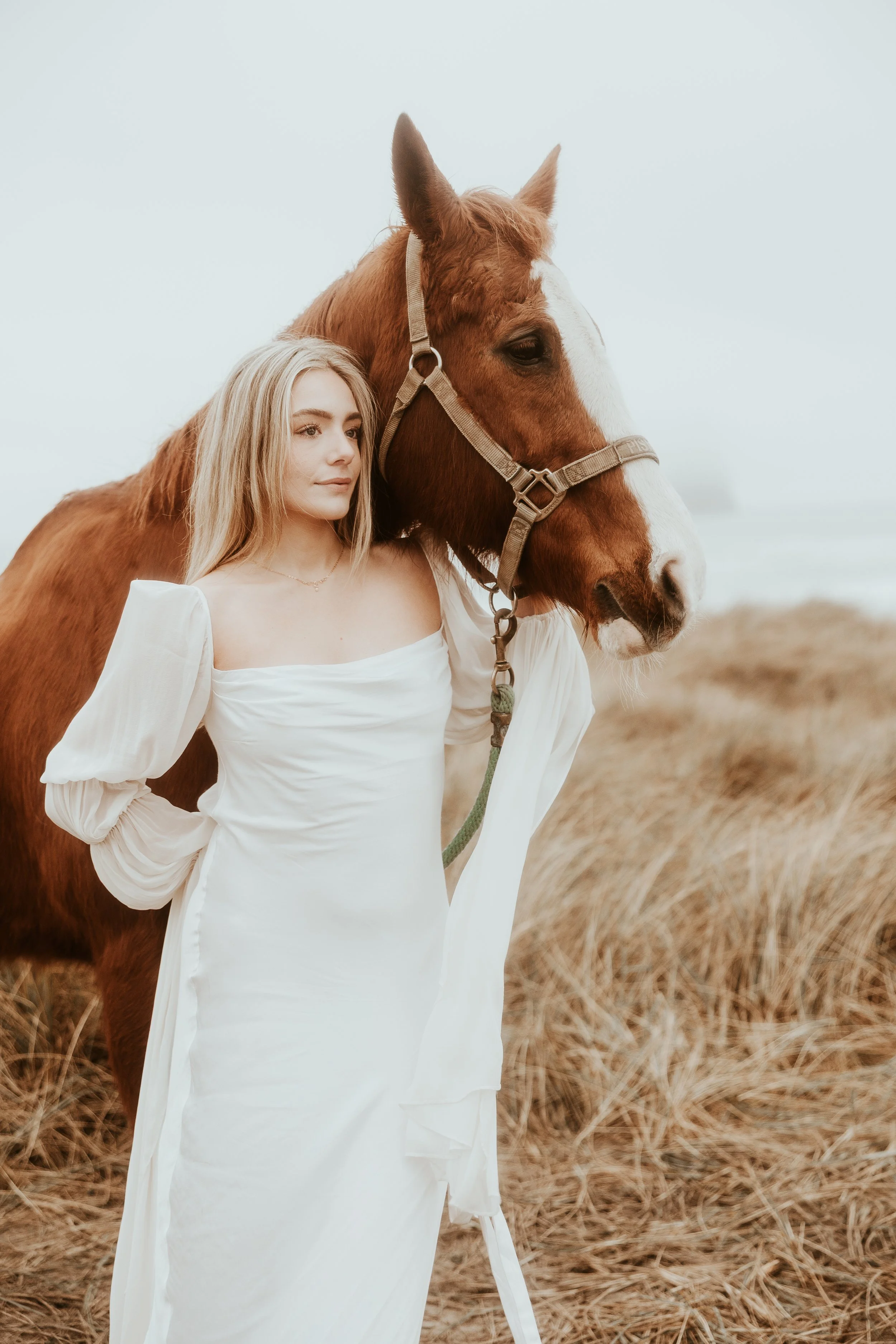 A young woman in a white dress standing beside a brown horse with a white blaze on its face in a field of dry grass near the coast.