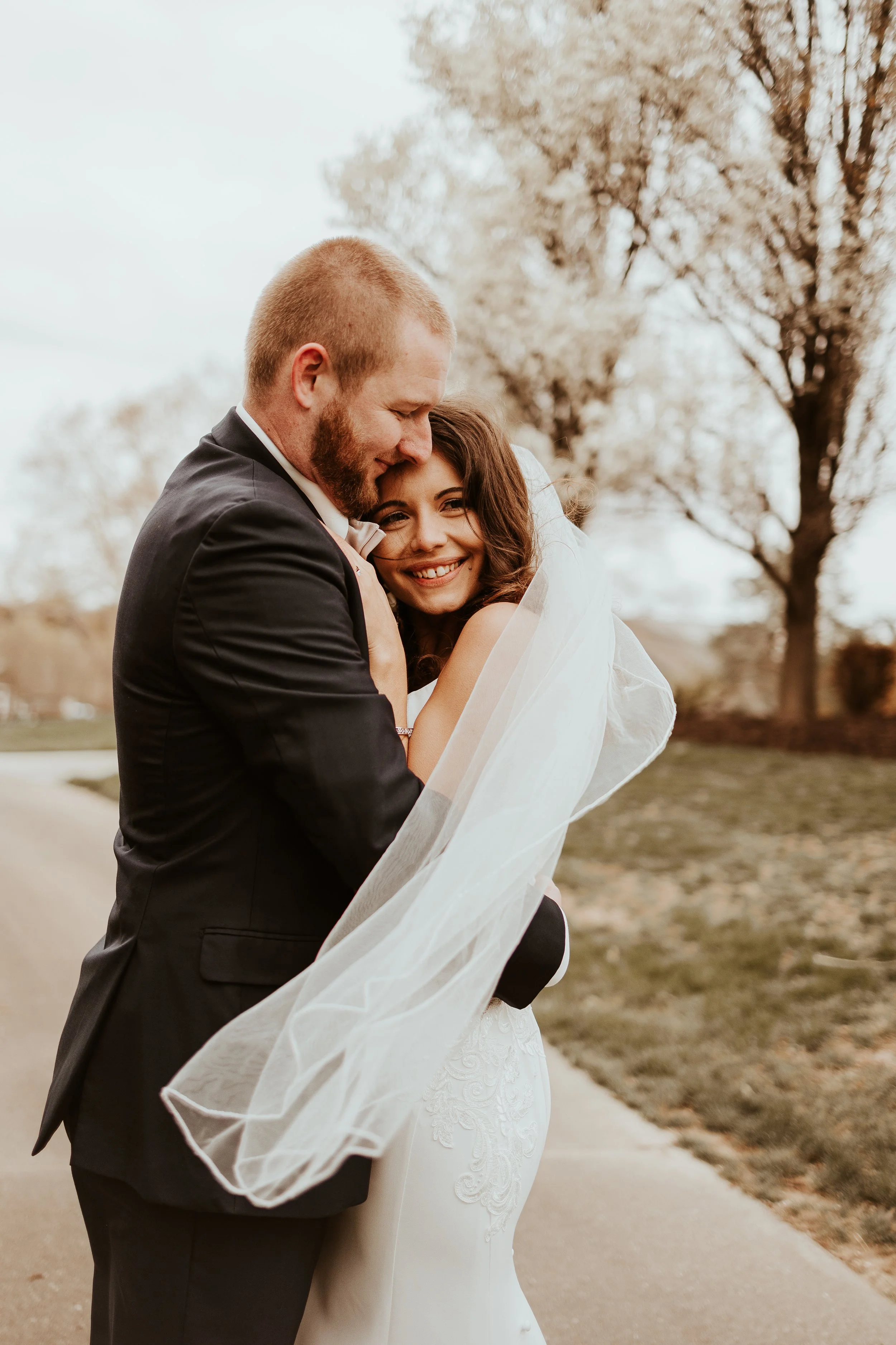 A newlywed couple hugging outdoors on a cloudy day, with bare trees in the background. The bride is smiling and wearing a white lace wedding gown and veil, while the groom is in a black suit.
