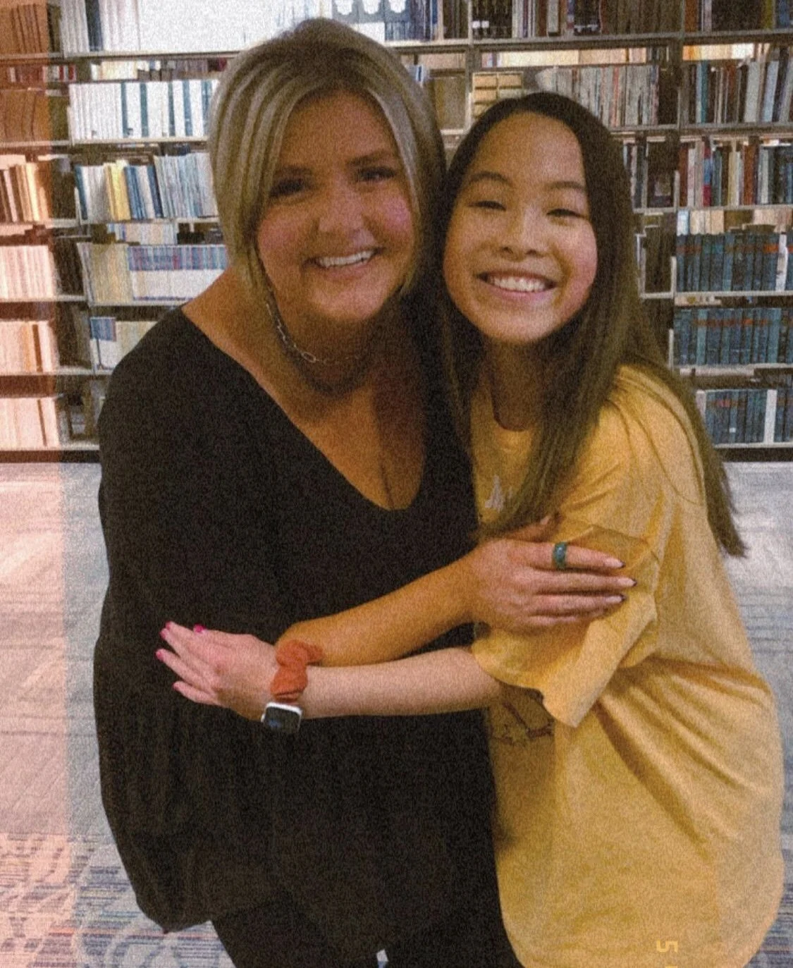 Two women hugging and smiling in a library with shelves of books in the background.