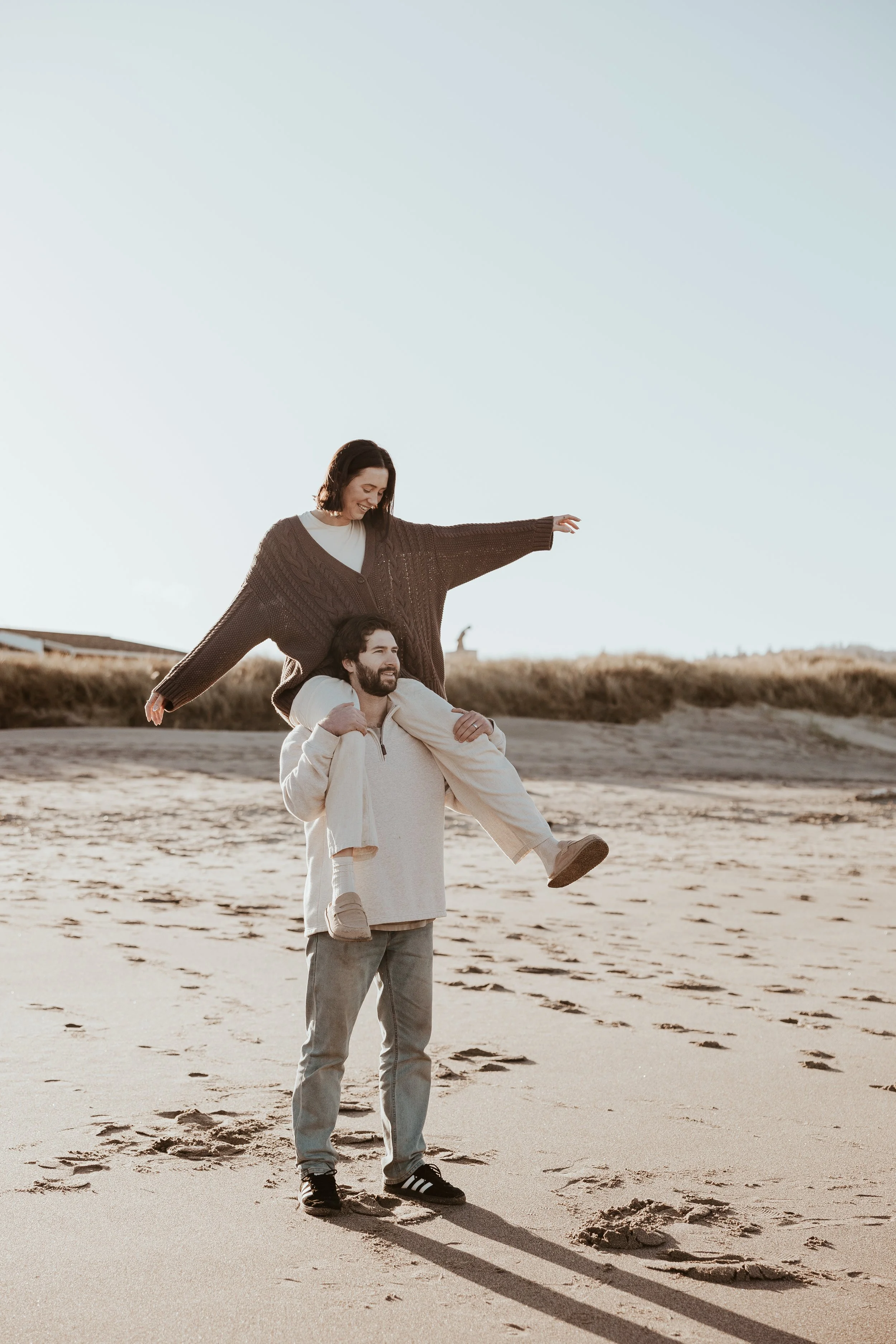 A man is carrying a woman on his shoulders on a sandy beach during sunset, both smiling and enjoying the moment.