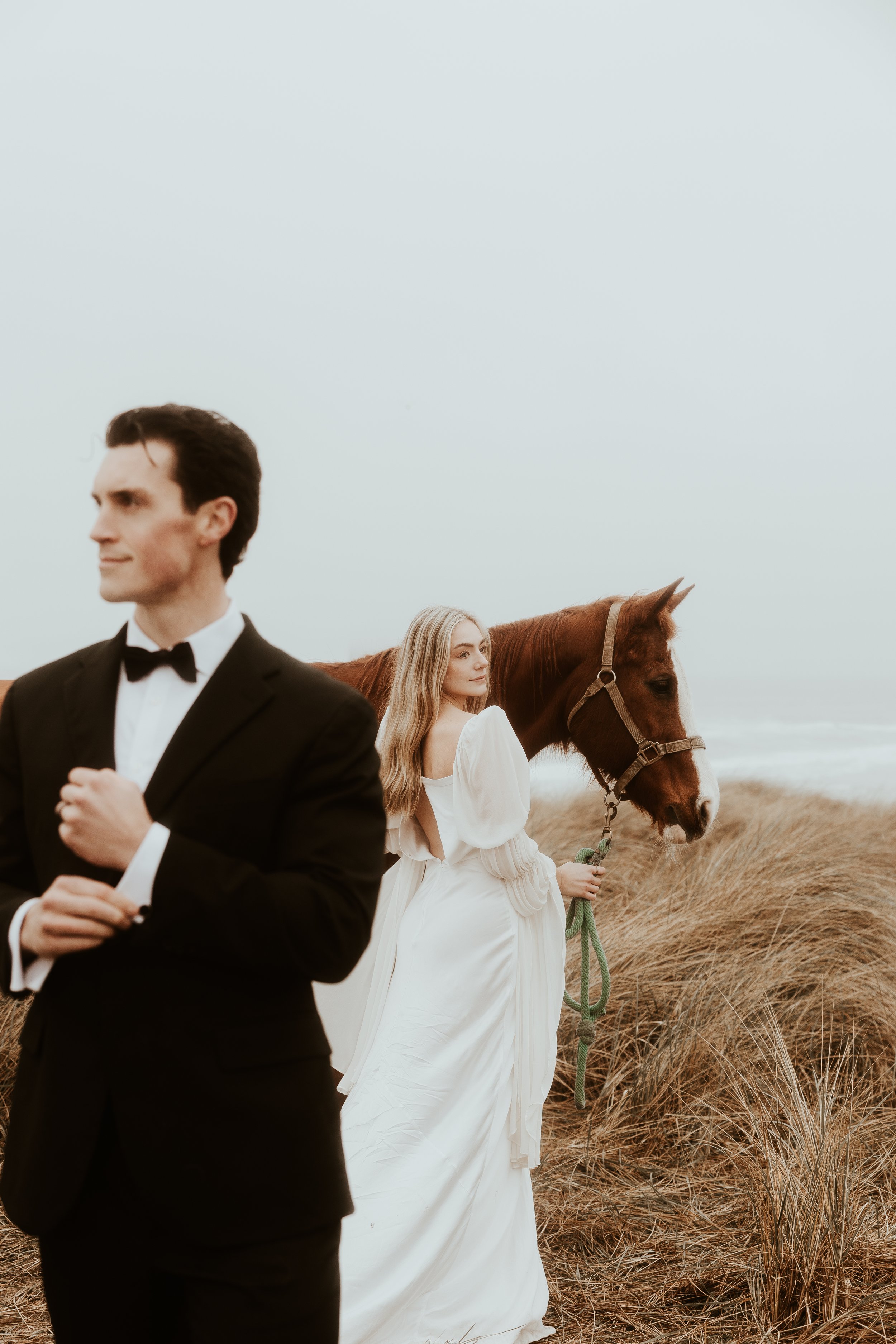 A man in a tuxedo standing in the foreground, and a woman in a white dress holding a horse's reins in the background on a grassy field near the ocean.