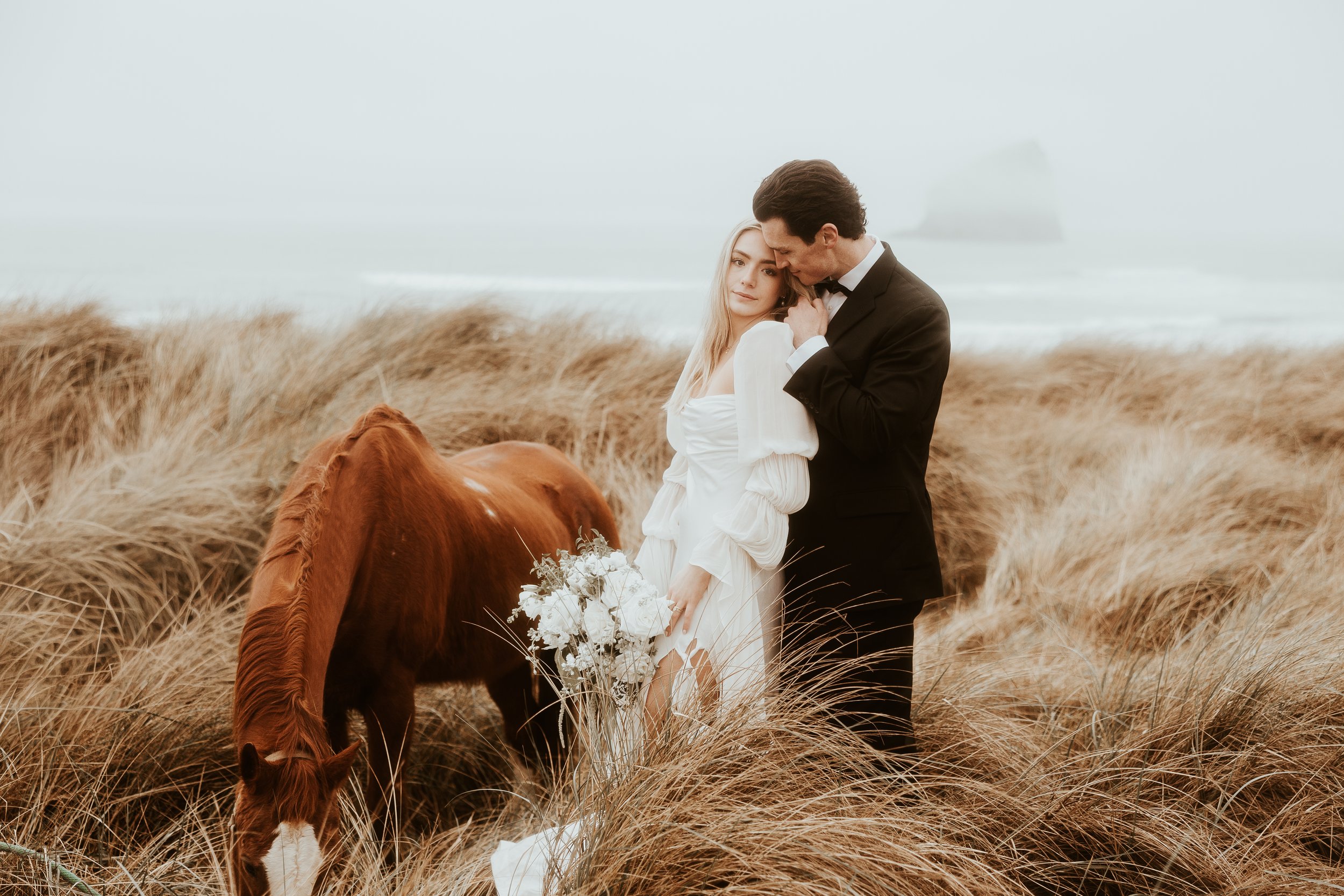 A couple in wedding attire standing in a field of tall grass near the ocean, with a horse grazing nearby. The bride holds a bouquet of white flowers; the groom leans in to kiss her on the forehead.
