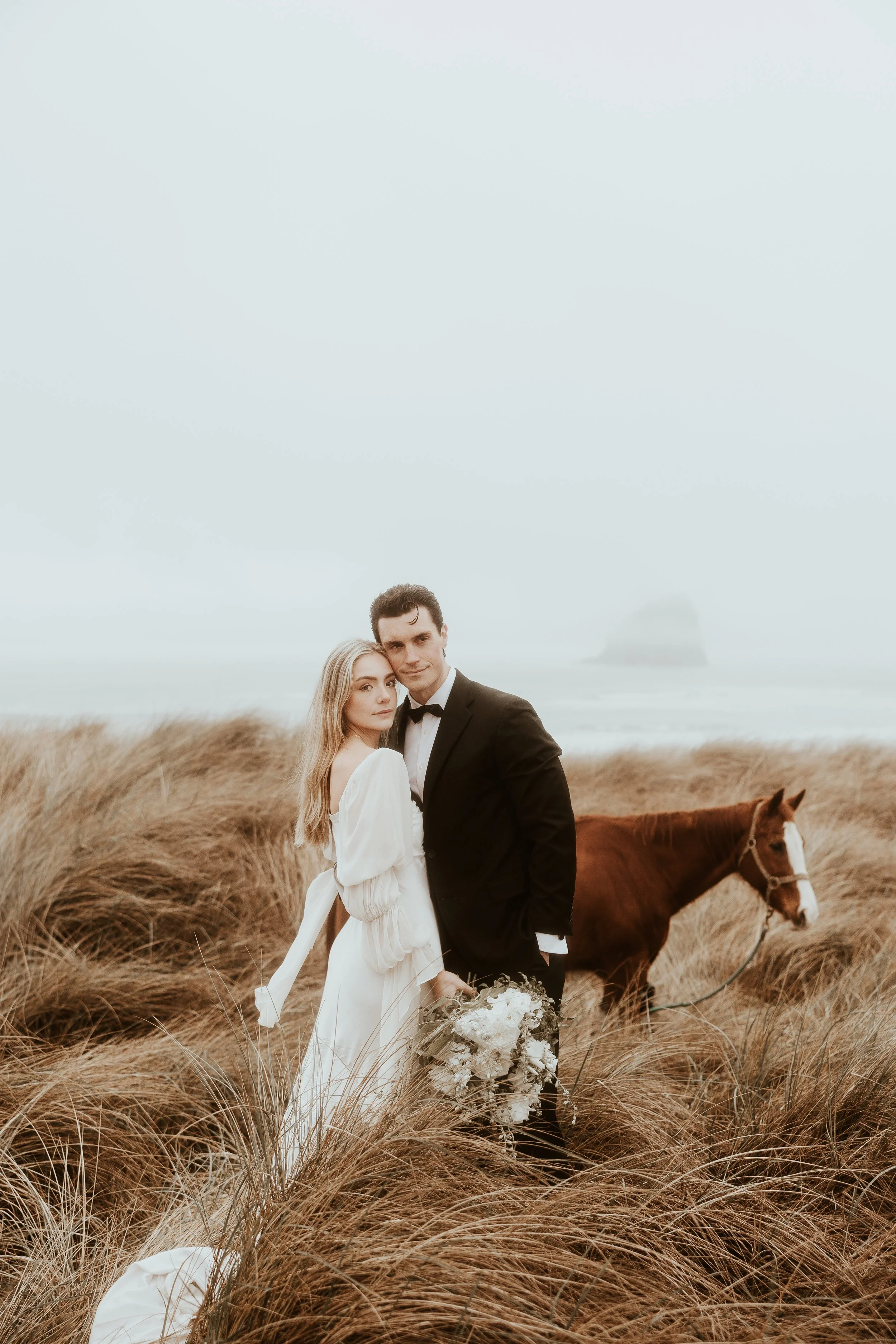 A bride and groom standing in a field of tall grass with a horse in the background, near the beach.
