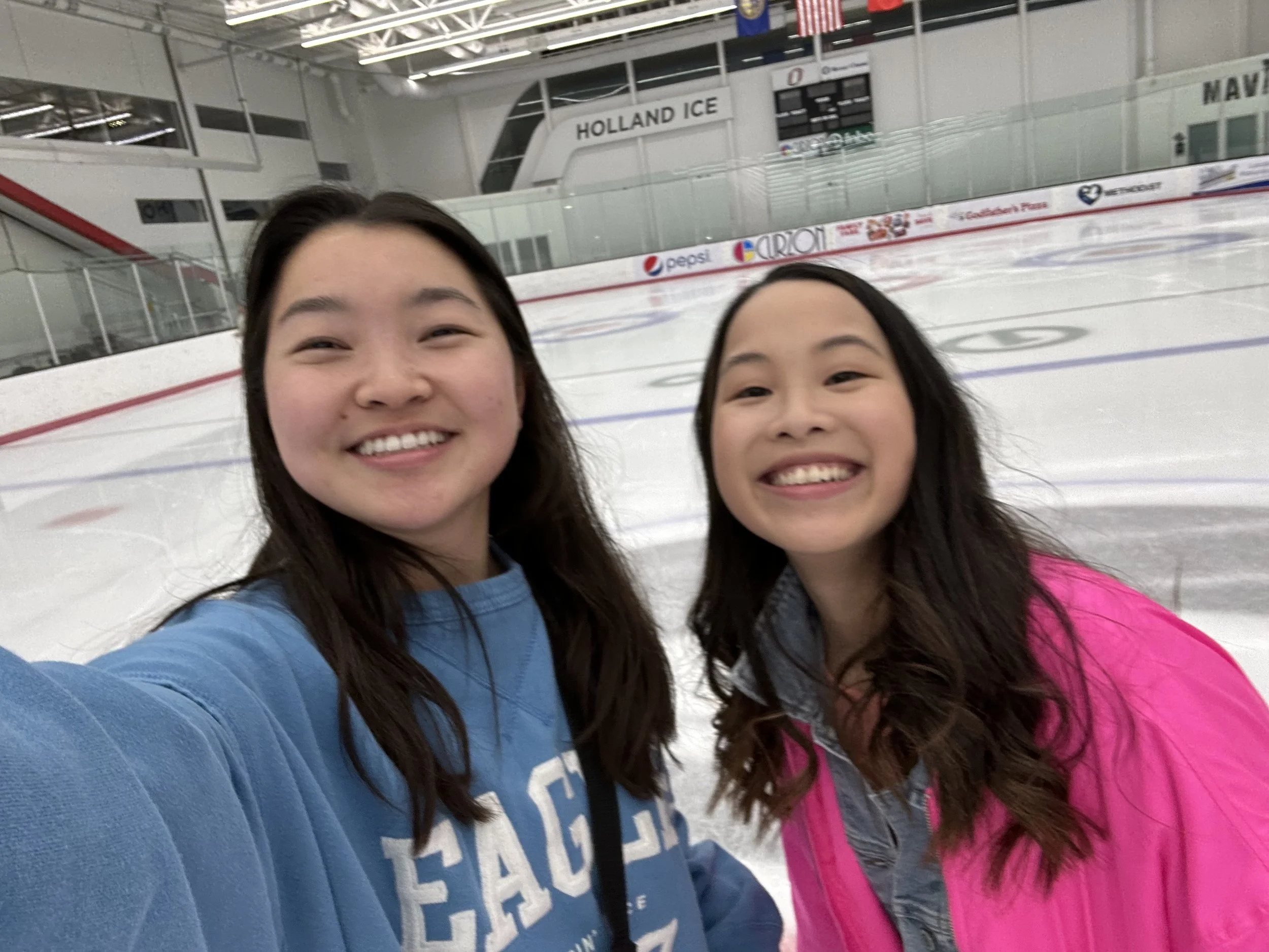 Two young women smiling on an indoor ice skating rink, with one wearing a blue sweatshirt and the other wearing a pink jacket.